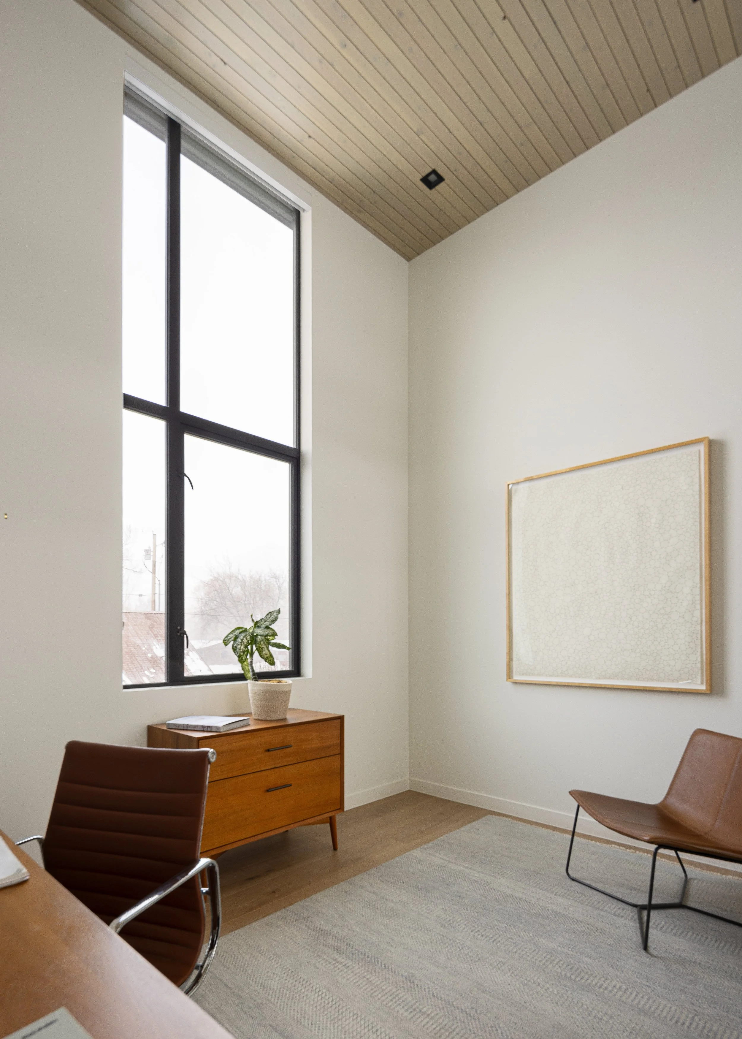 Office at Snow King in Jackson, Wyoming. Modern office room with wood ceiling, large window, potted plant, wooden cabinet, framed art, and brown chairs.