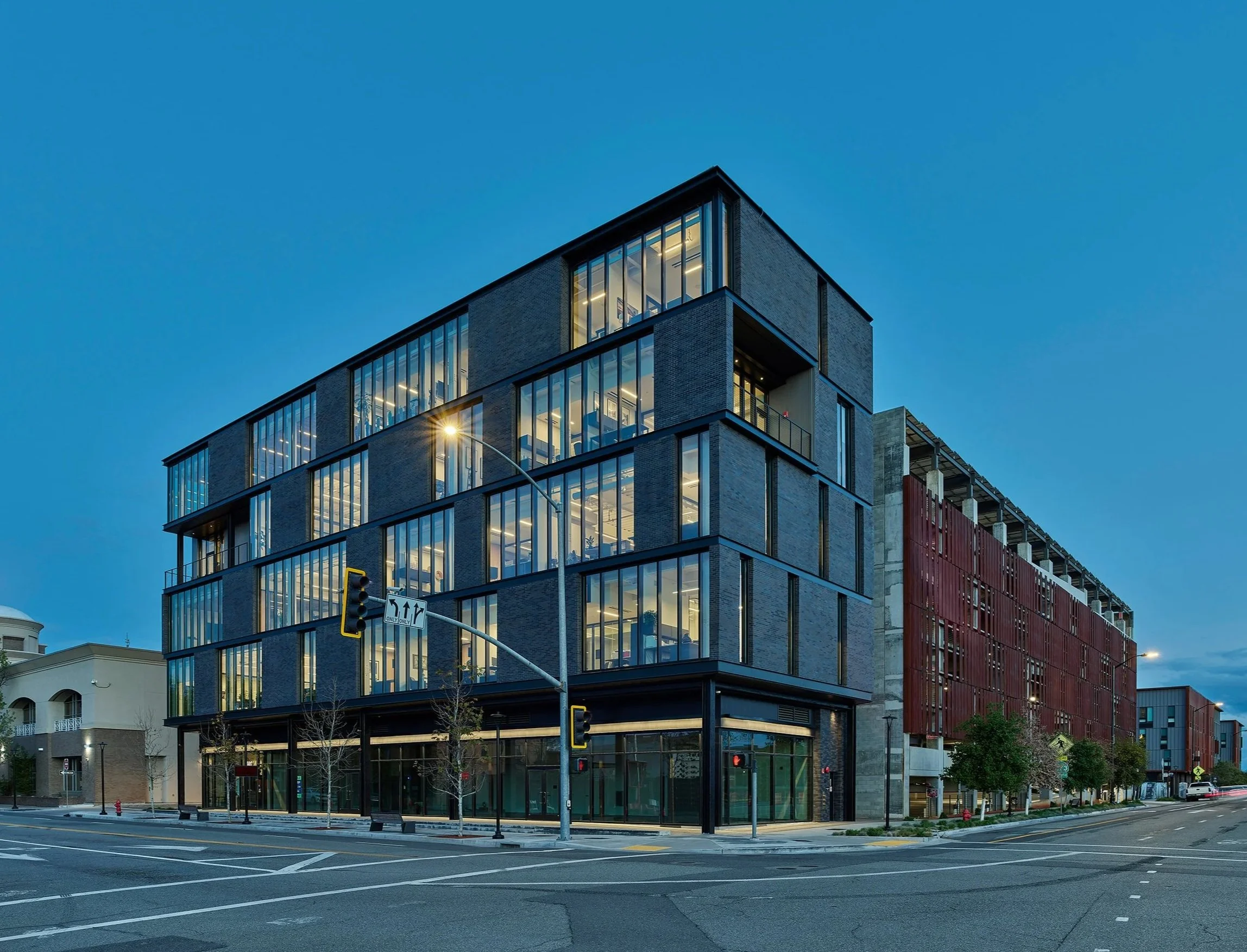 Modern multi-story building with large glass windows on a city street during dusk, with streetlights and traffic signals visible.