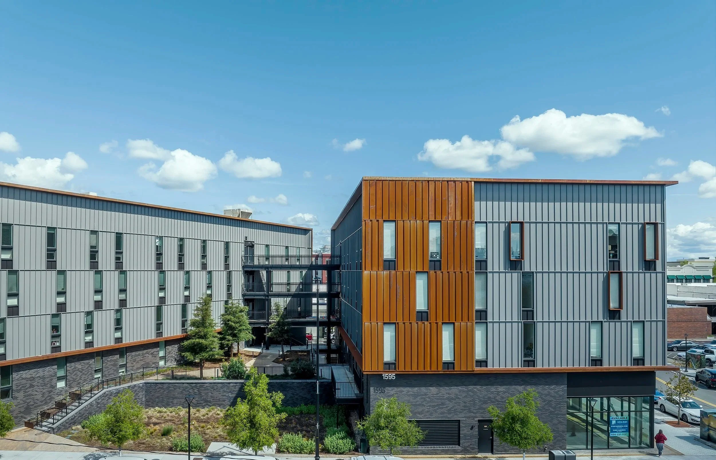 Block 7 Development in Redding, California. California Place affordable housing. Modern multi-story buildings with mixed metal and wood facades, with some trees and a landscaped area in the foreground.