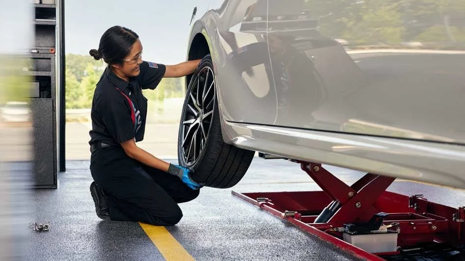 A woman kneeling next to a grey car, inspecting the front wheel, in a garage or service center.