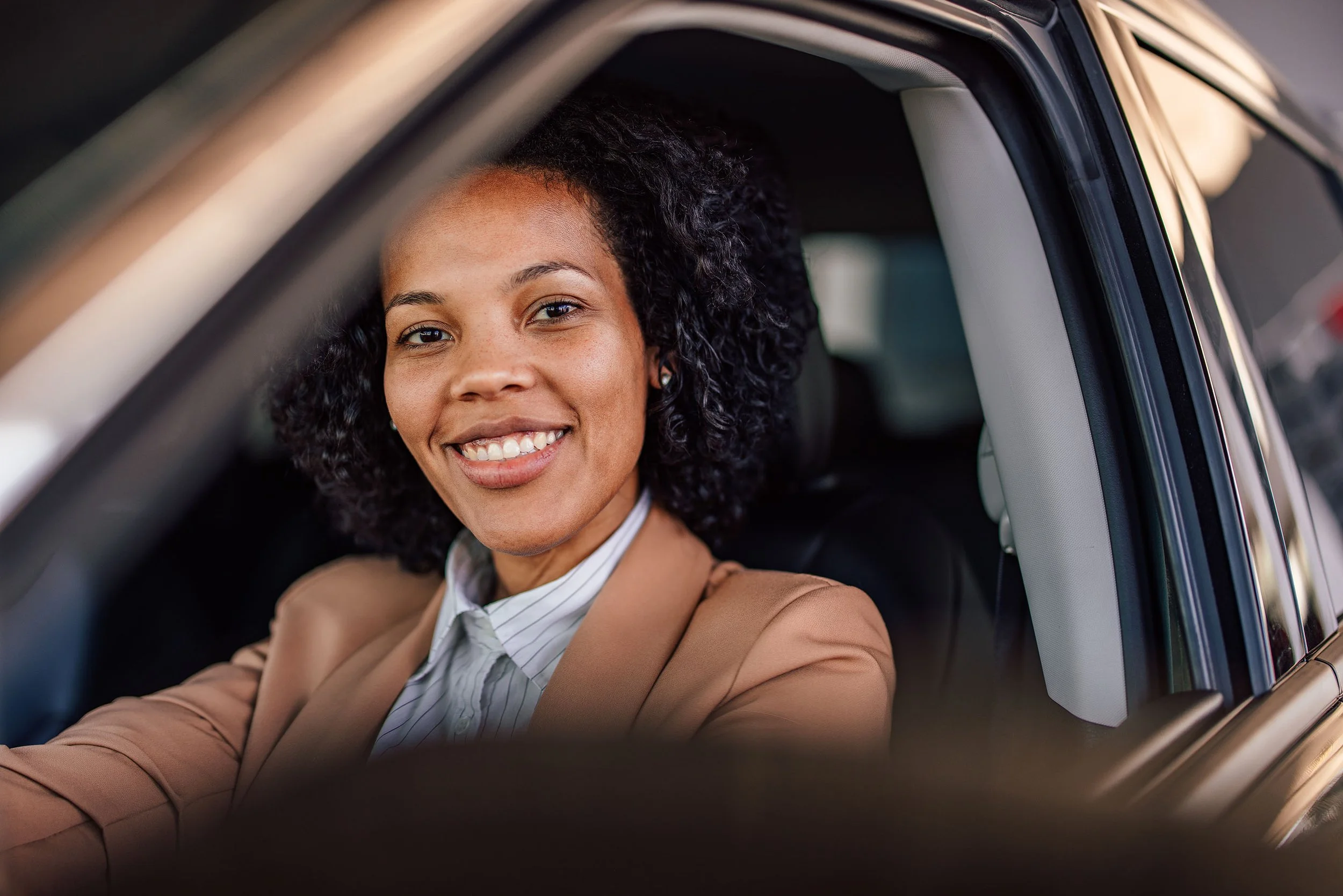 A woman with curly hair smiling, sitting in the driver's seat of a car, wearing a tan blazer and a collared shirt.