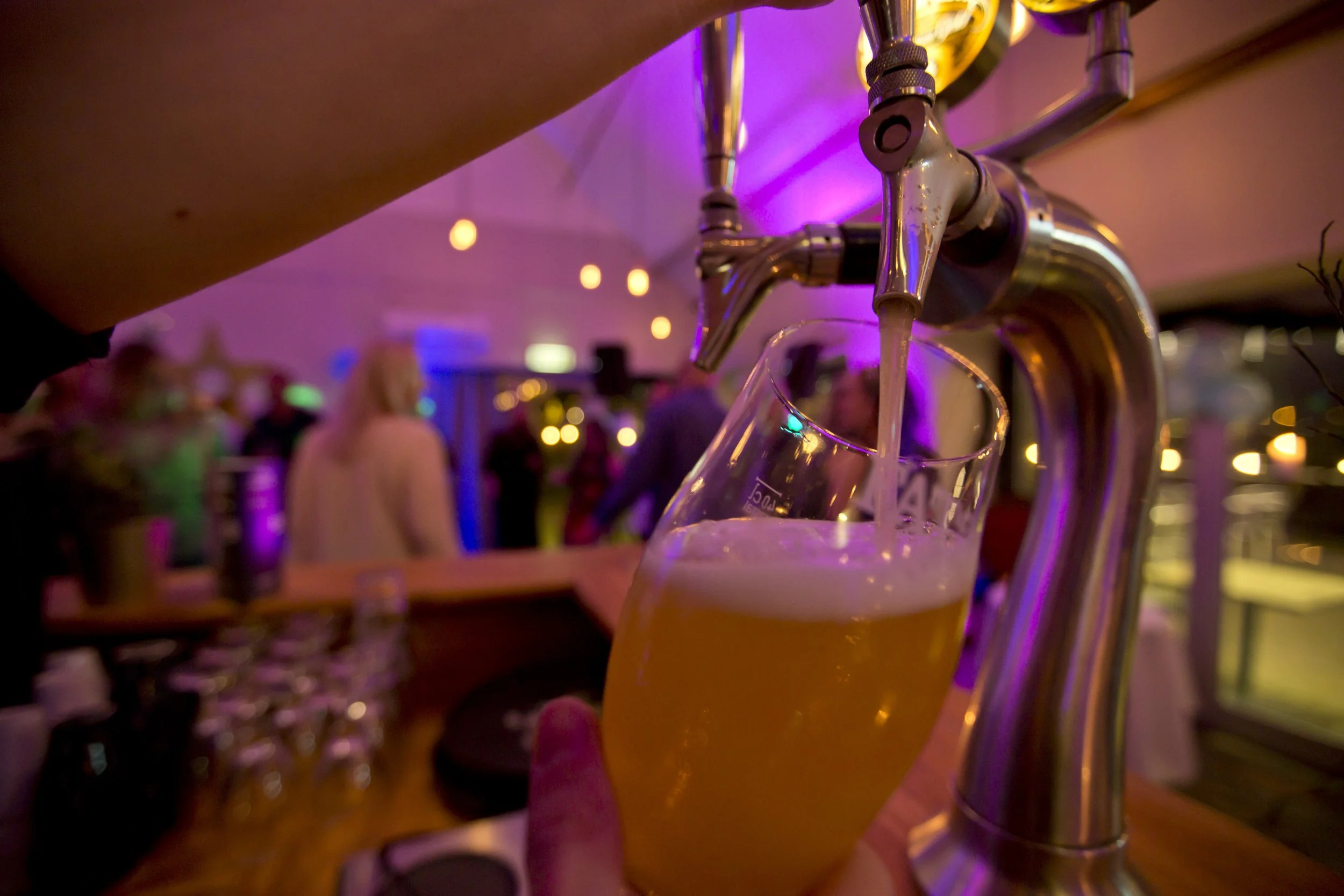 A glass of beer being poured from a tap in a lively bar or nightclub with purple lighting and blurred patrons in the background.
