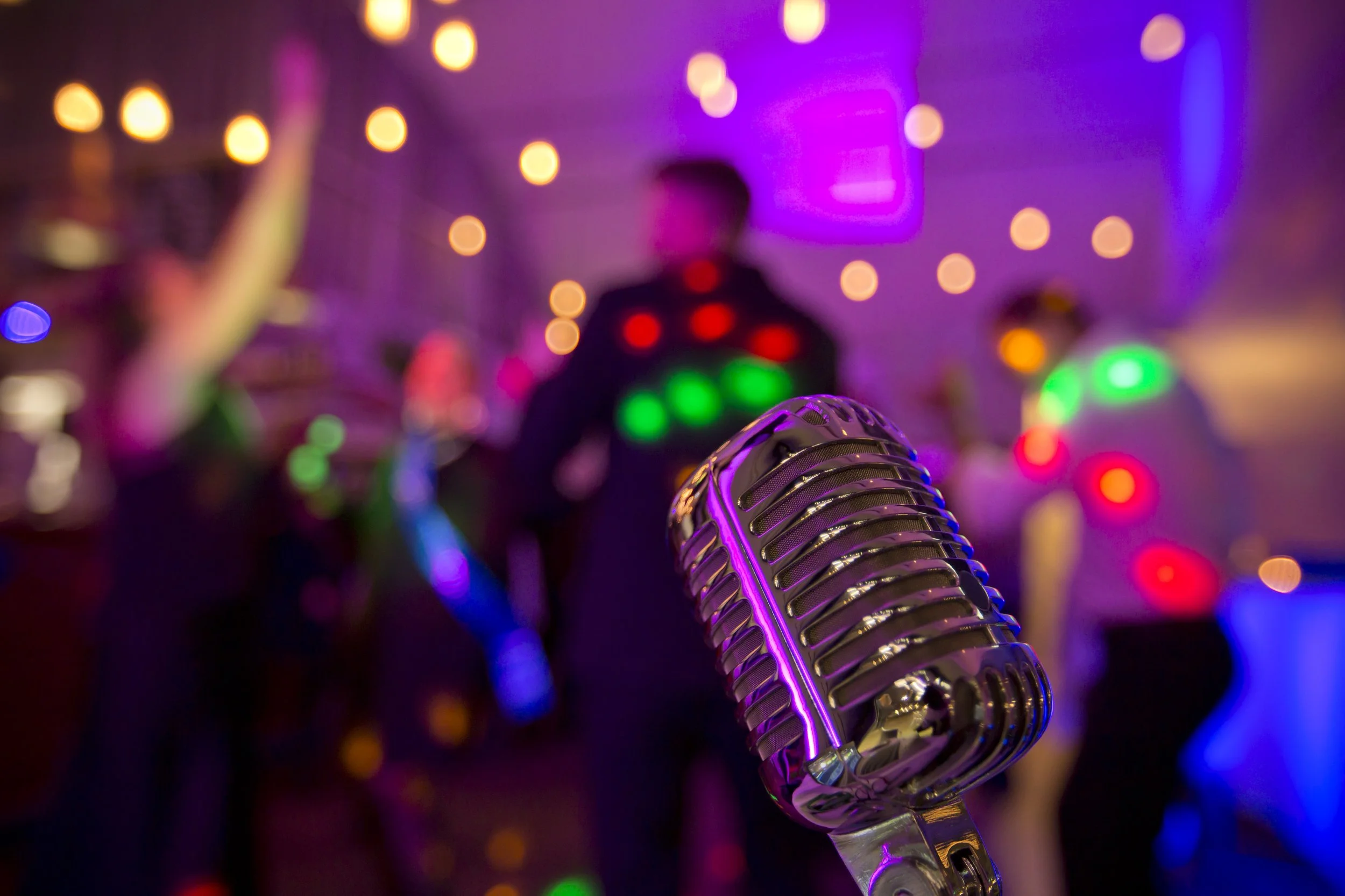 A close-up of a vintage microphone on a stand with a backdrop of a blurry party scene with people dancing and colorful lights.