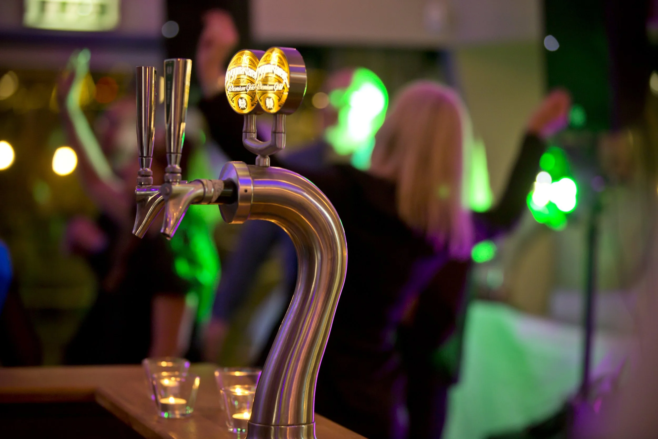 Close-up of a beer tap in a bar with colorful lights in the background, with people socializing.