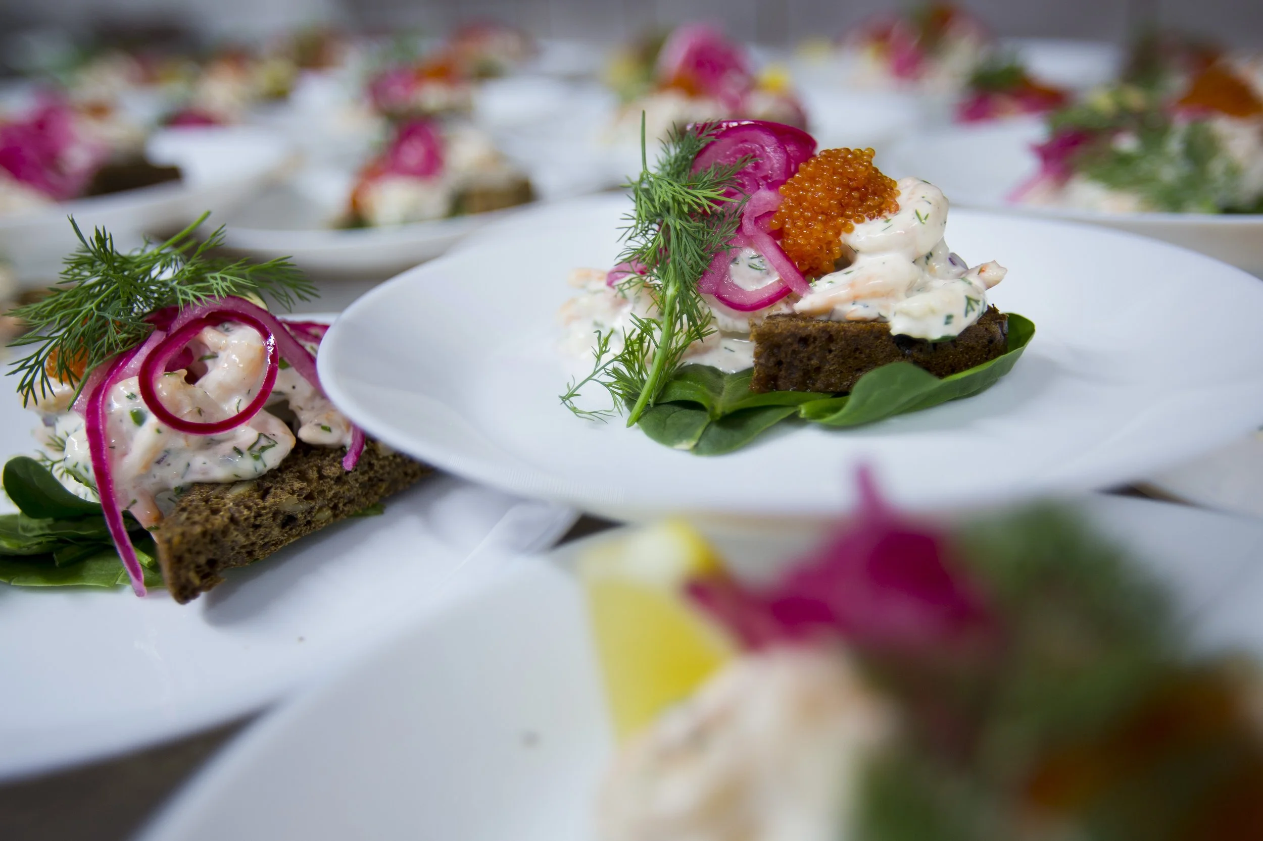 Appetizer of dark rye bread topped with creamy white spread, pickled red onions, fresh dill, and orange fish roe, served on a white plate with a green leaf underneath.