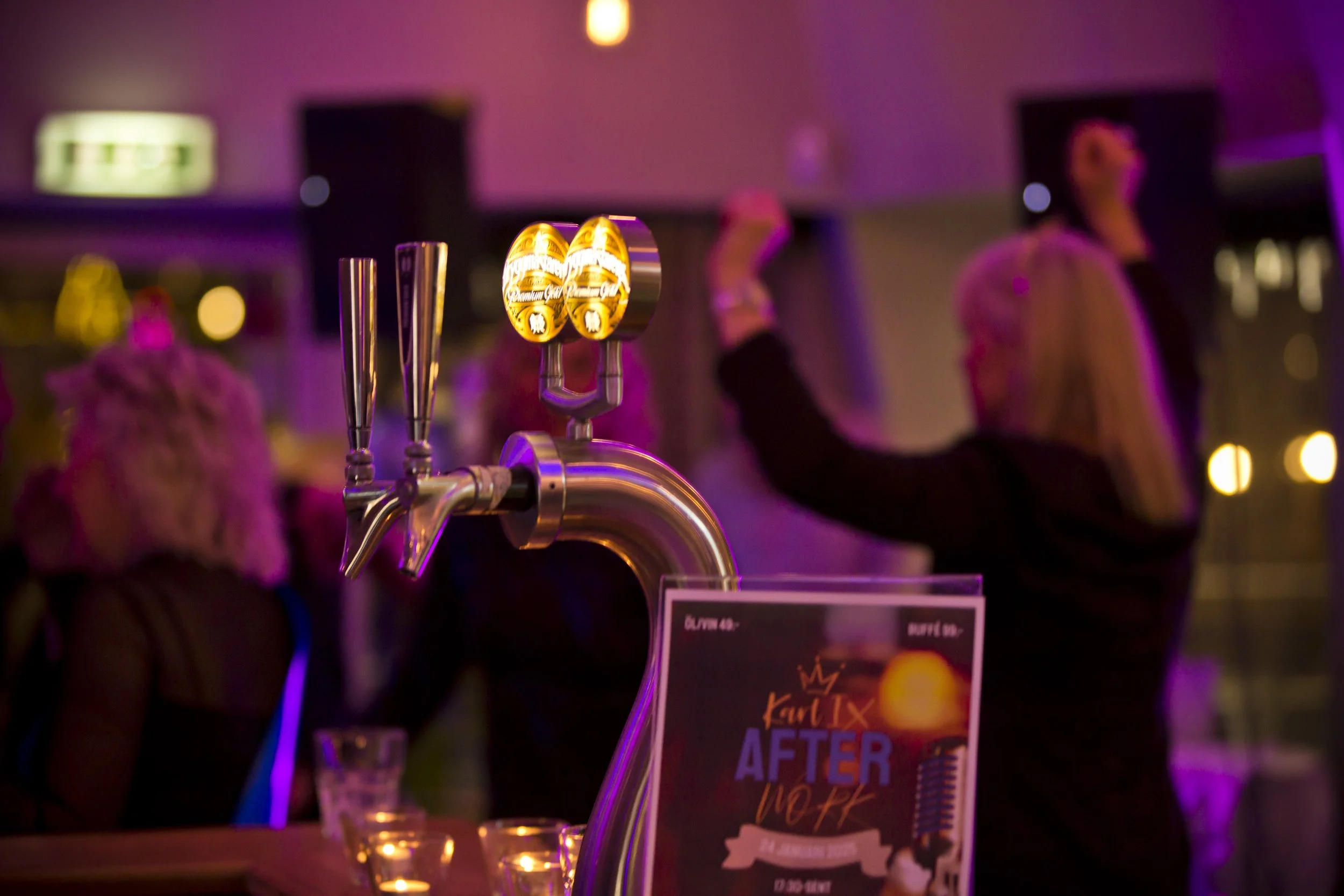 A bar tap with two beer spouts illuminated by purple and yellow lighting, with a blurred woman in the background raising her glass in a social setting.