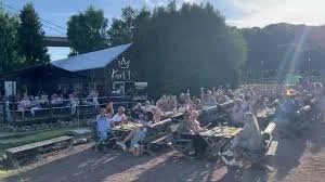 People sitting at outdoor picnic tables near train tracks with trees and a building in the background
