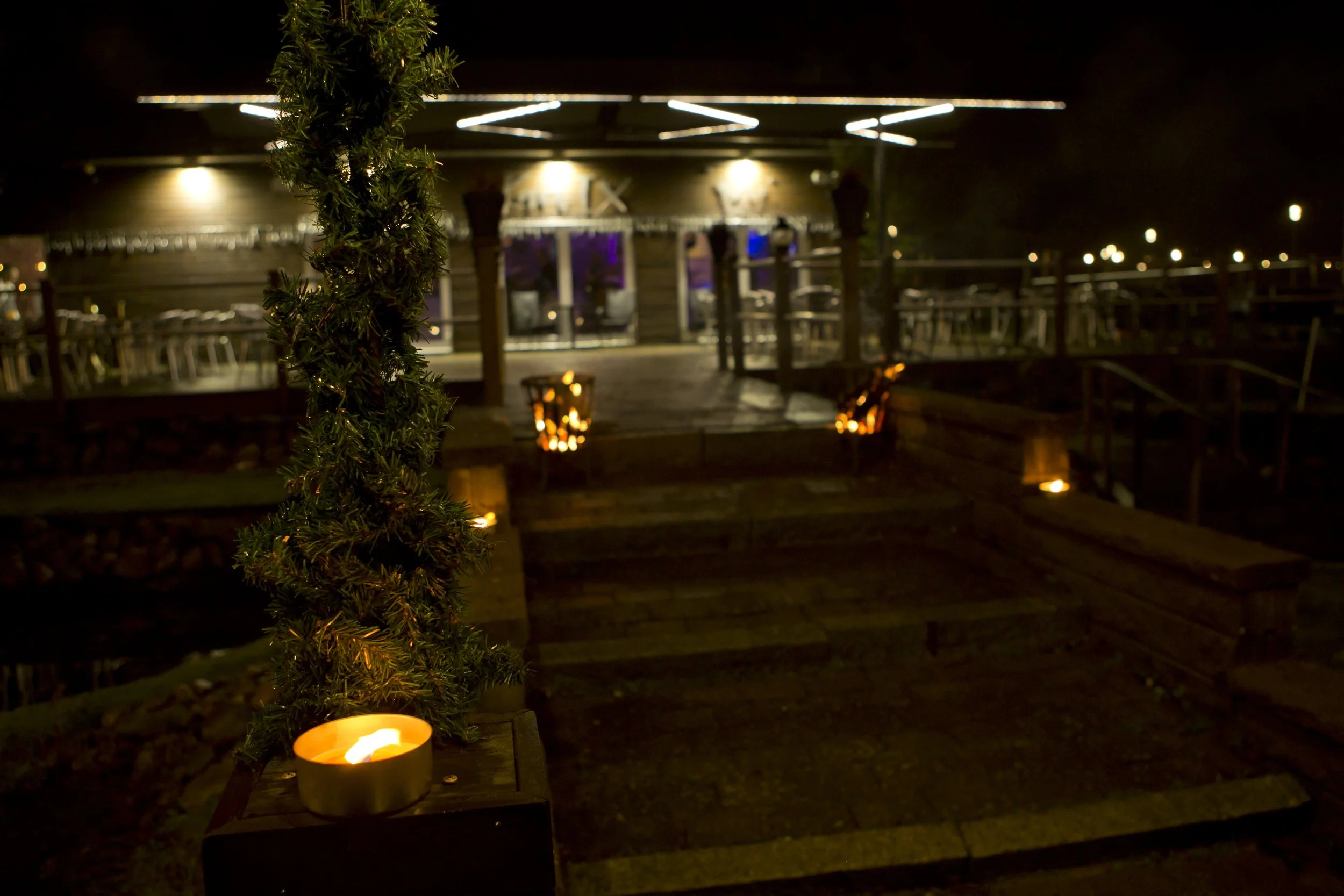 Nighttime scene of an outdoor patio with a wooden deck, decorated with candles and a small wrapped Christmas tree, leading to a lit building with string lights.