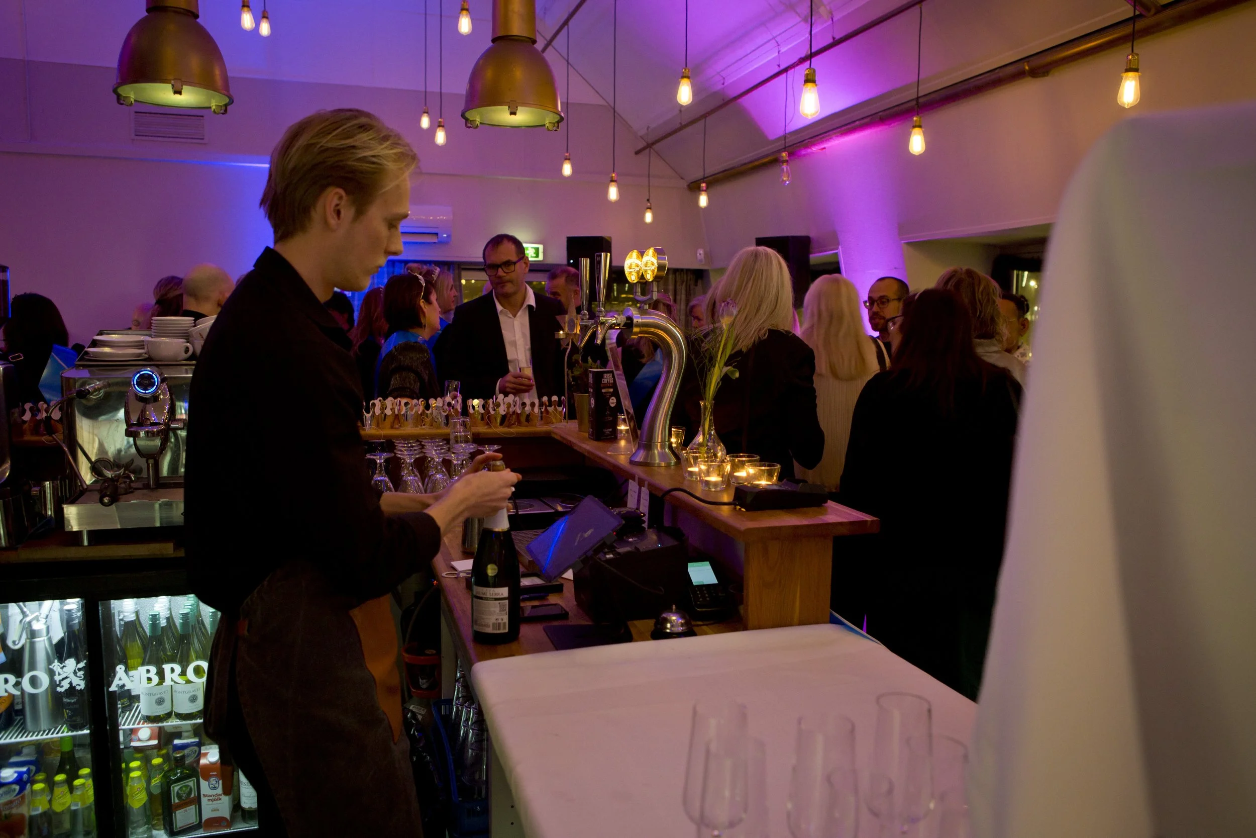 A bar filled with people socializing, illuminated with warm and pink lighting, with a bartender preparing drinks, and a refrigerator stocked with beverages in the foreground.