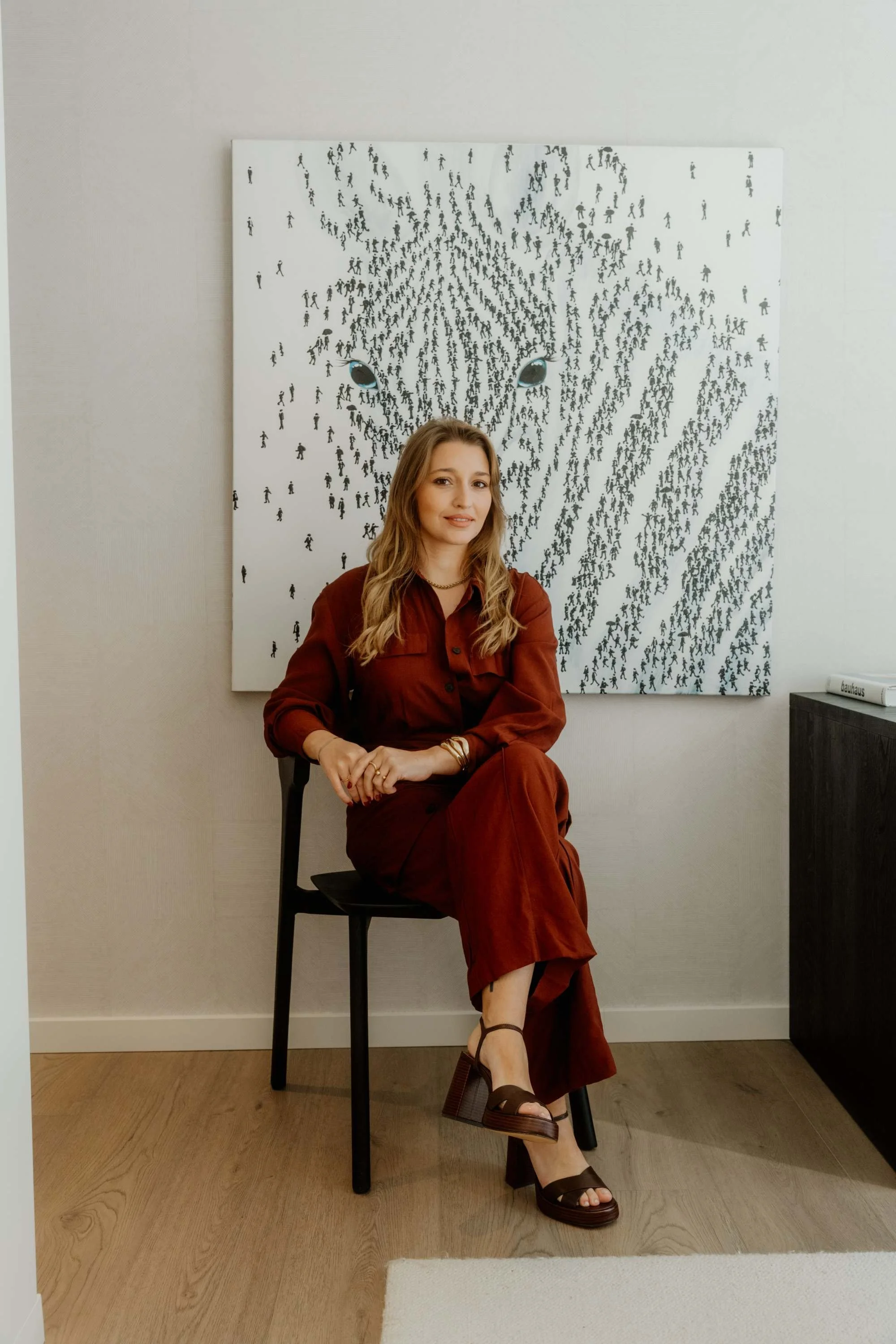 A woman in a rust-colored outfit sitting on a black chair in front of artwork featuring a tiger face made up of small black silhouettes of people.