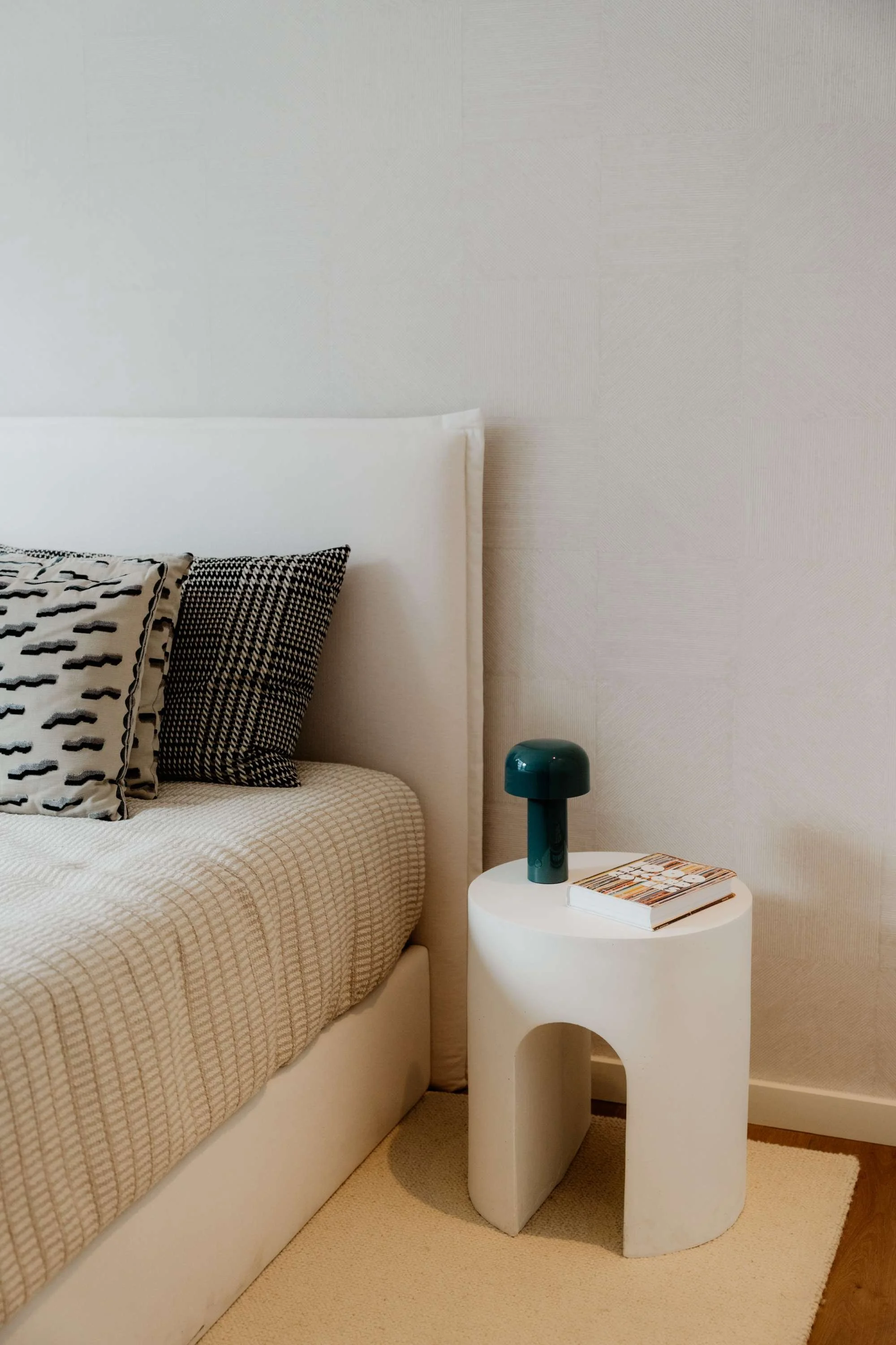 A bedroom with a white bed, black and white patterned pillows, a white side table with a dark green lamp, a magazine, a beige carpet, and a white wall.