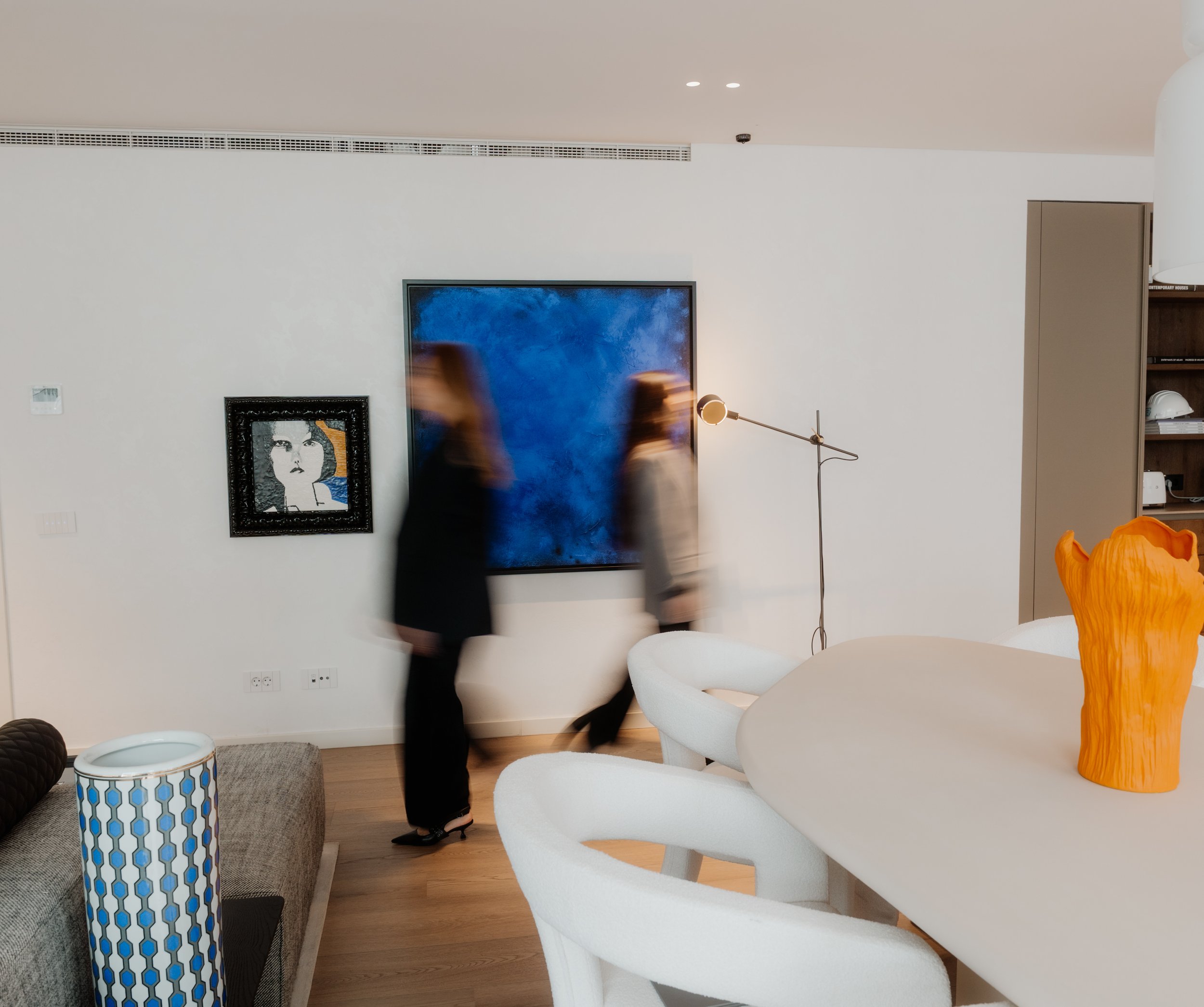 Modern living room with white walls, wall art depicting a woman's face, a large blue abstract painting, a floor lamp, and white chairs. A blurred person appears to be walking.