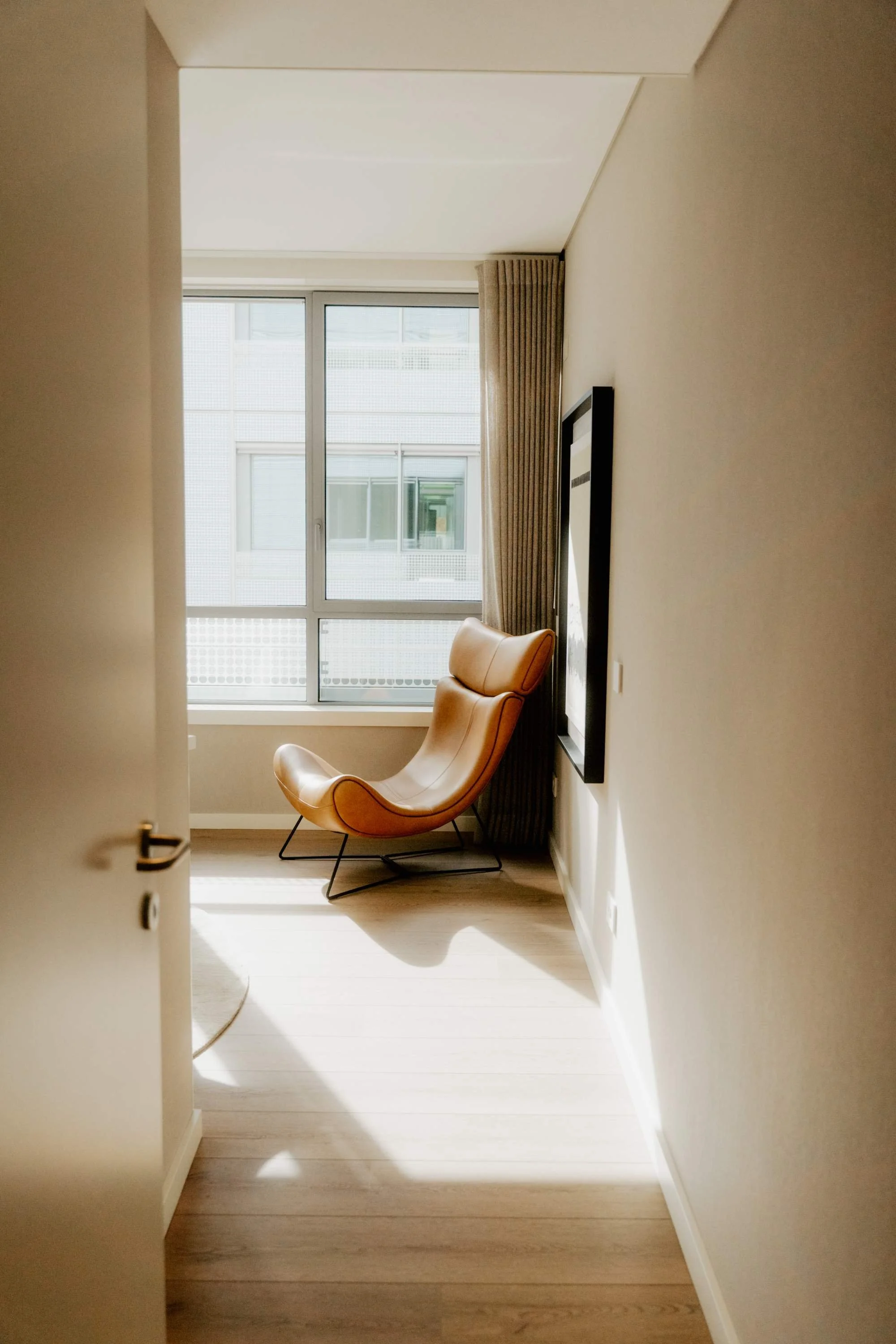 Interior of a room with a leather lounge chair by the window, sunlight casting shadows on the wooden floor, framed art on the wall, and beige curtains.