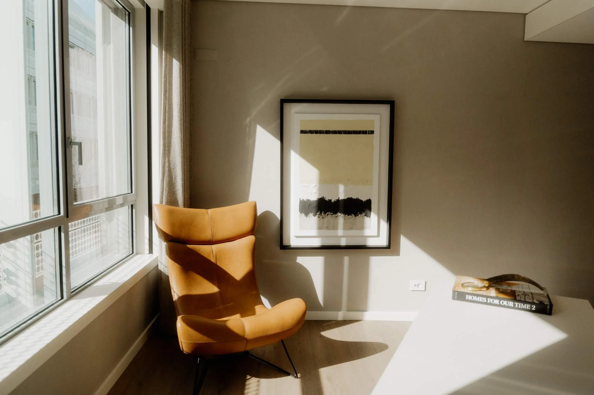 Interior of a room with a large window, a tan armchair, framed abstract art on the wall, and a white table with a book titled 'HOMES FOR OUR TIME 2' and a decorative object, illuminated by sunlight creating shadows.