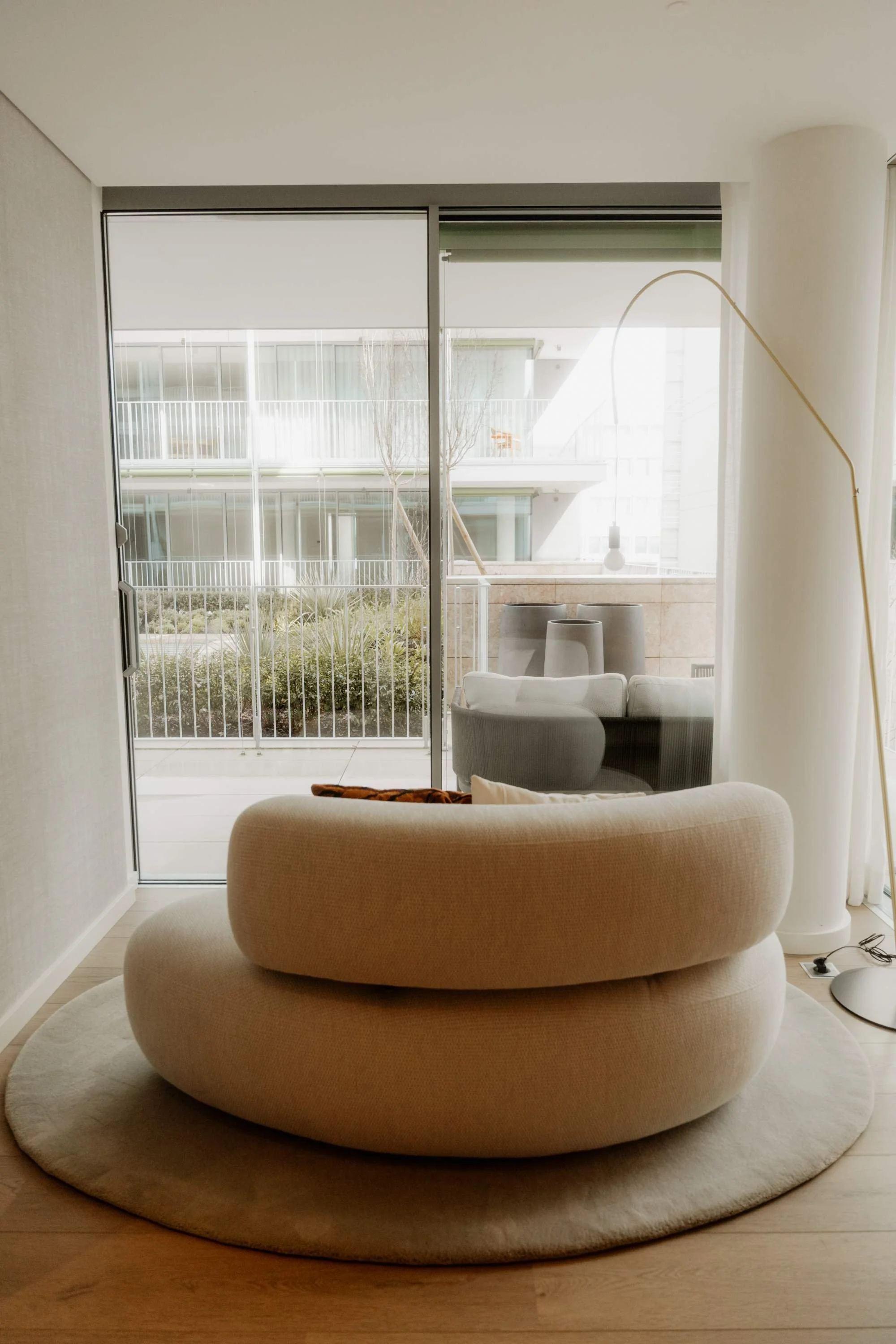 Living room with curved beige sofa, glass sliding door leading to a balcony with outdoor furniture, and tall white floor lamp.