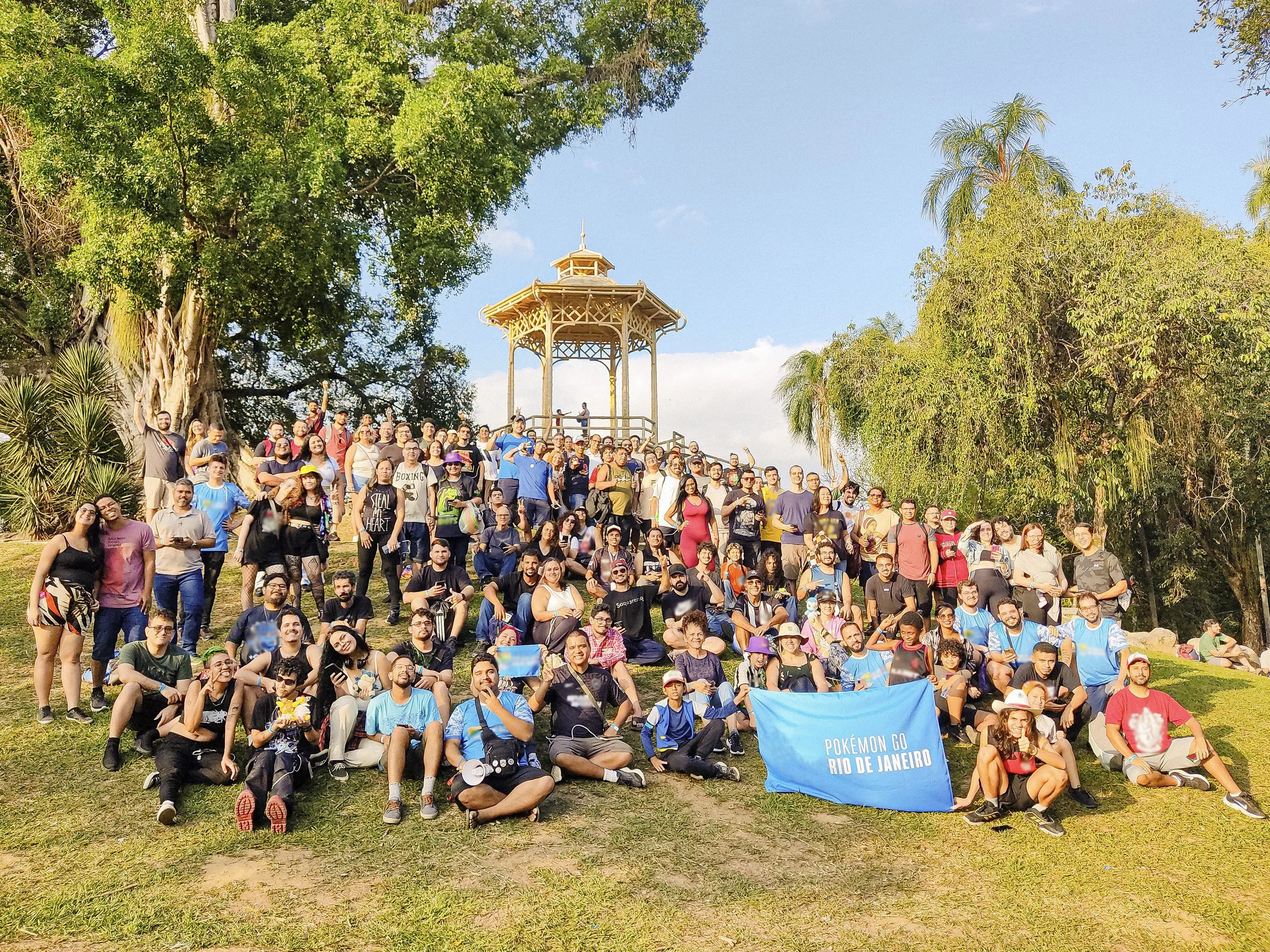 Um grande grupo de pessoas reunido ao ar livre em uma colina gramada com árvores e um coreto em estilo colonial ao fundo, segurando uma faixa azul com os dizeres 'Pokémon GO Rio de Janeiro'.