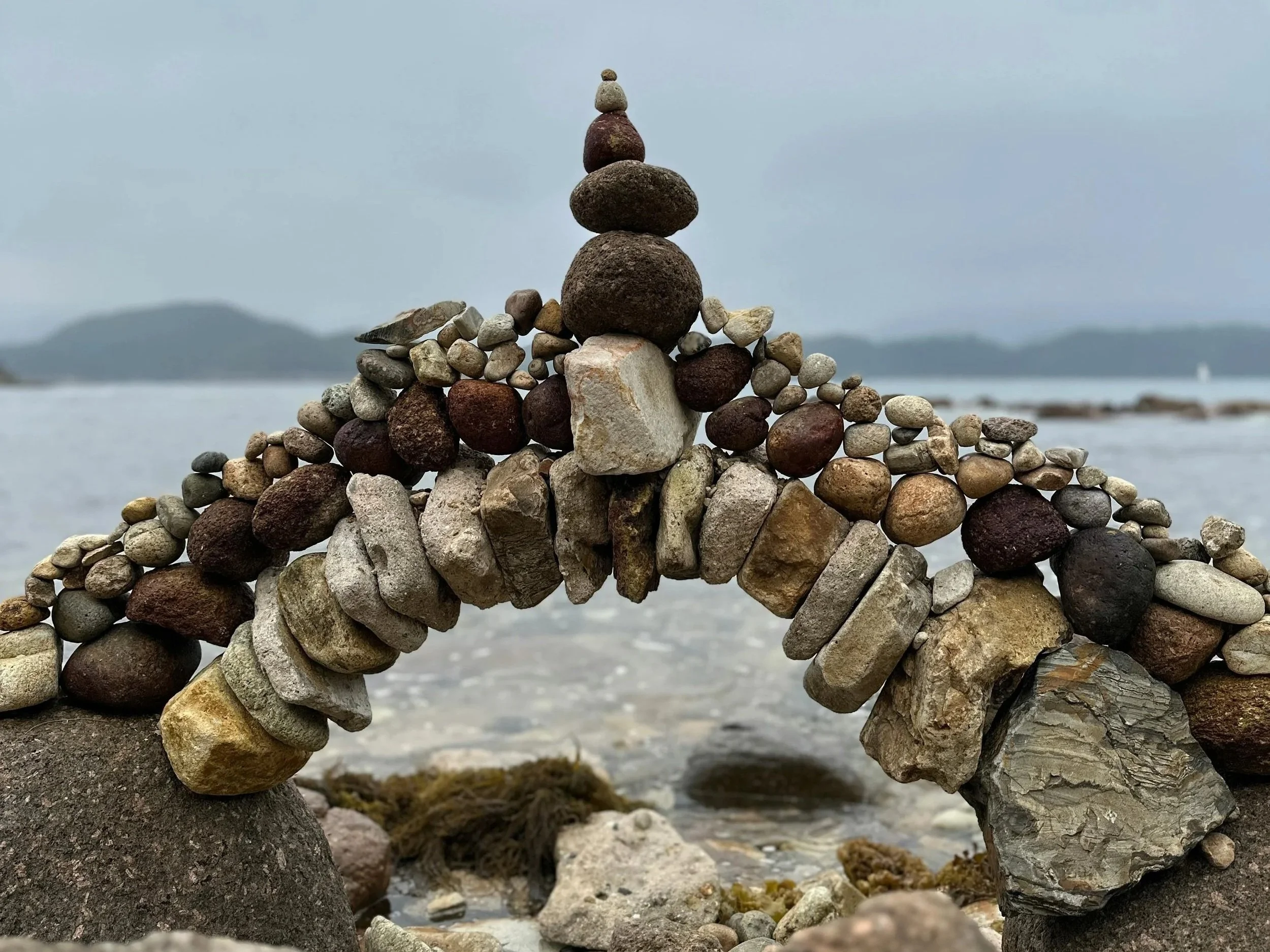 A rock sculpture of a whale made from balanced stones on a beach, with water and distant islands in the background.