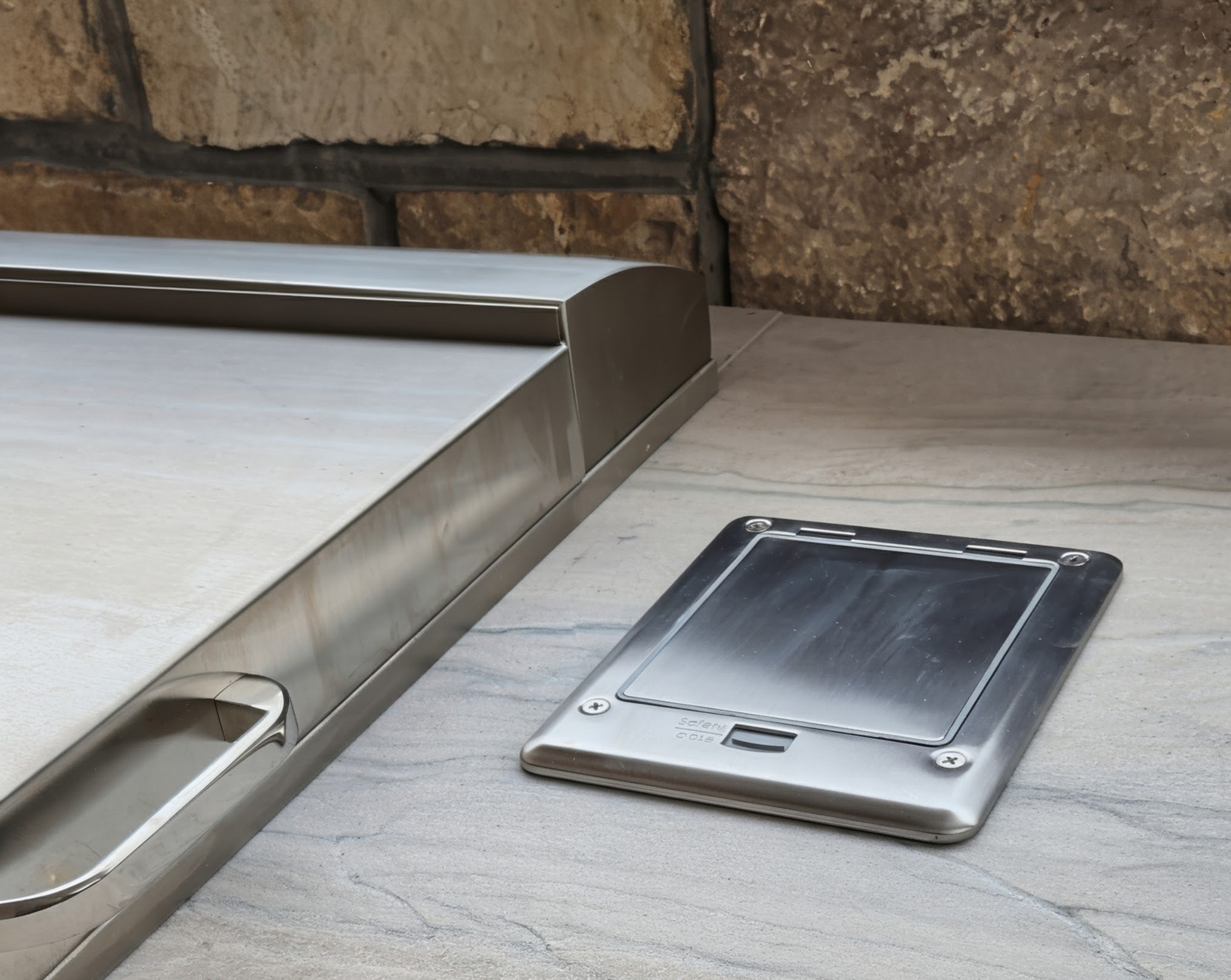 Close-up of a stainless steel sneeze guard and a metal plate on a countertop against a stone wall.