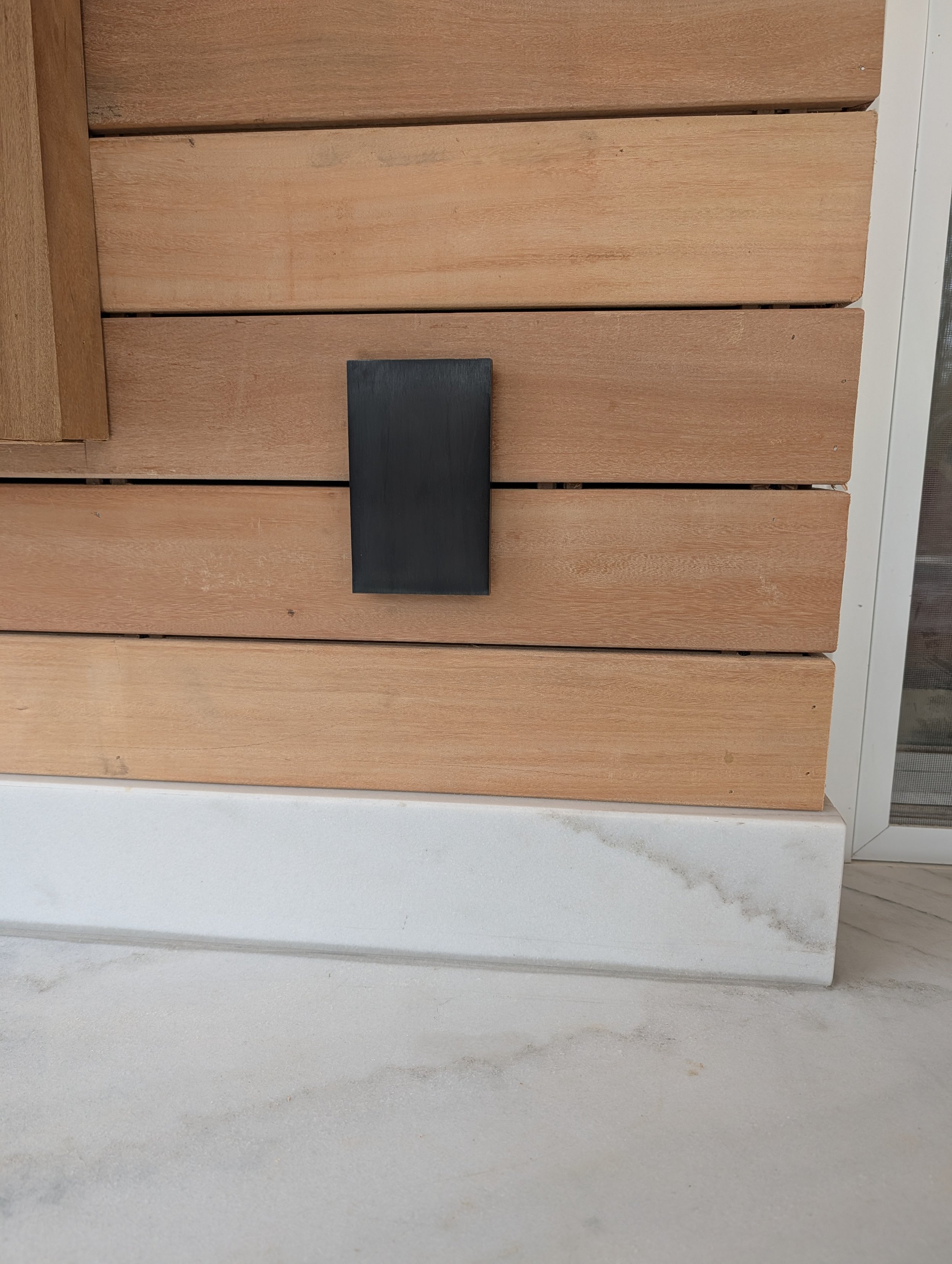 Close-up of a wooden wall with horizontal planks and a black rectangular object attached to it, with a white base beneath and part of a glass door visible on the right.