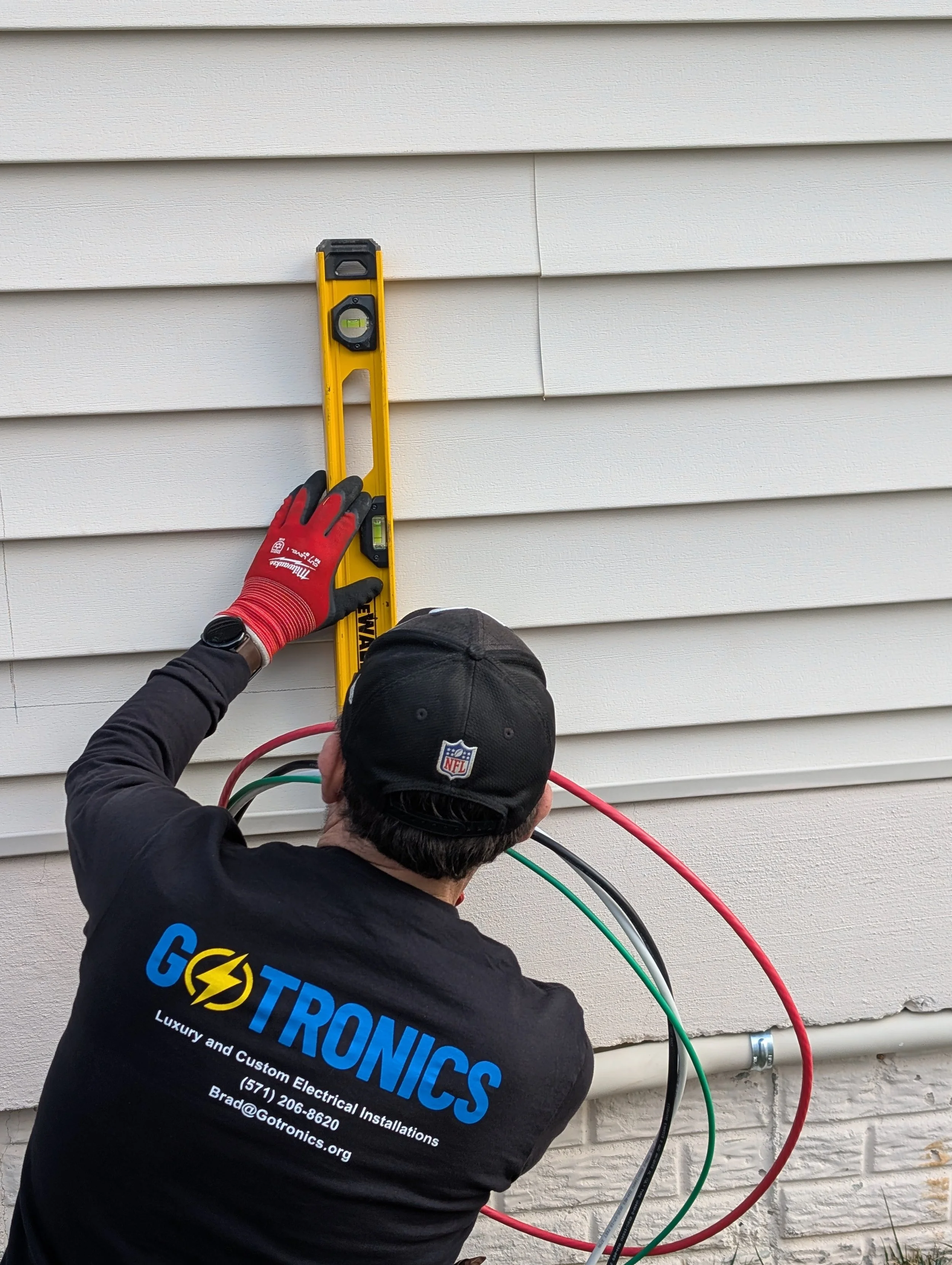A technician with a GOTORICS shirt is using a yellow level to measure a house's exterior wall during electrical work.
