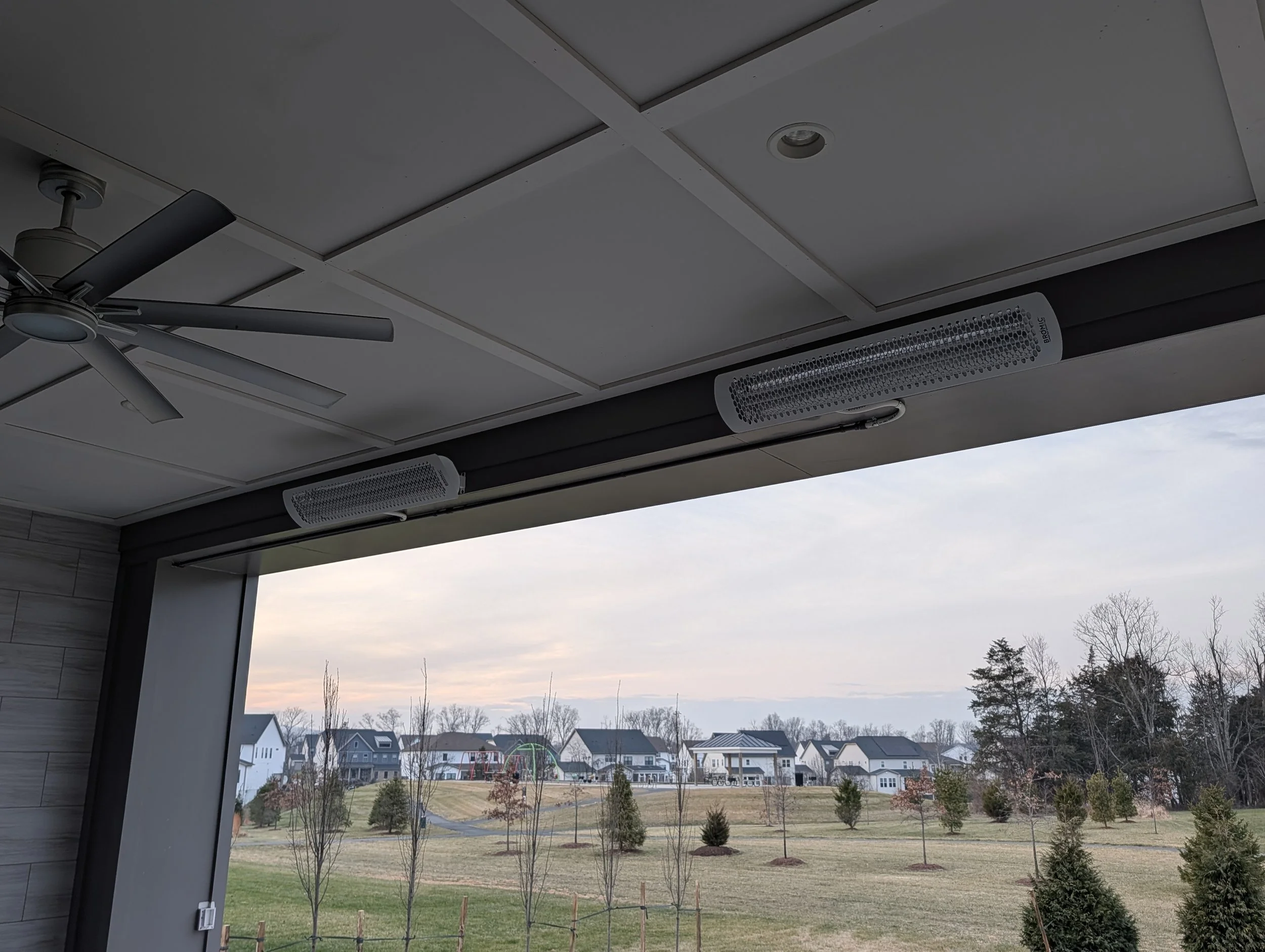 View from inside a building's covered porch or balcony showing a ceiling fan, mounted electric heaters, and an outdoor landscape with grass, trees, and residential houses under a cloudy sky.