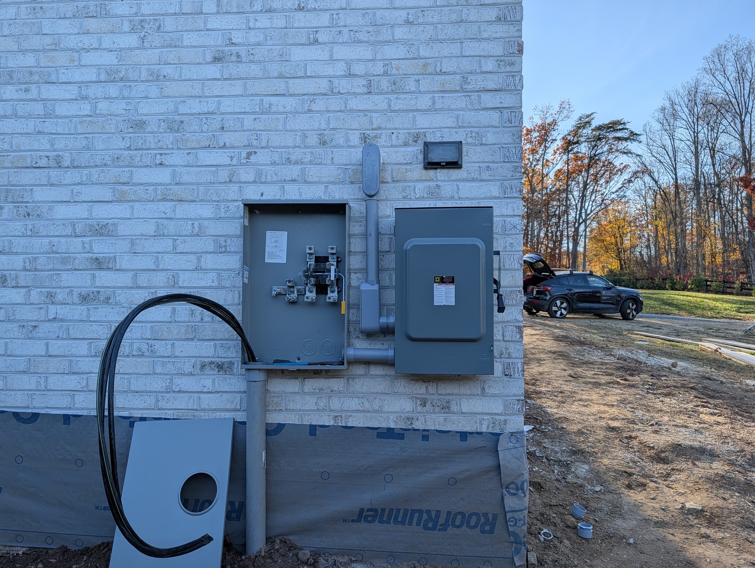 Electrical meter and panel on a brick exterior wall of a building under construction, with construction site and parked car in the background during daylight.