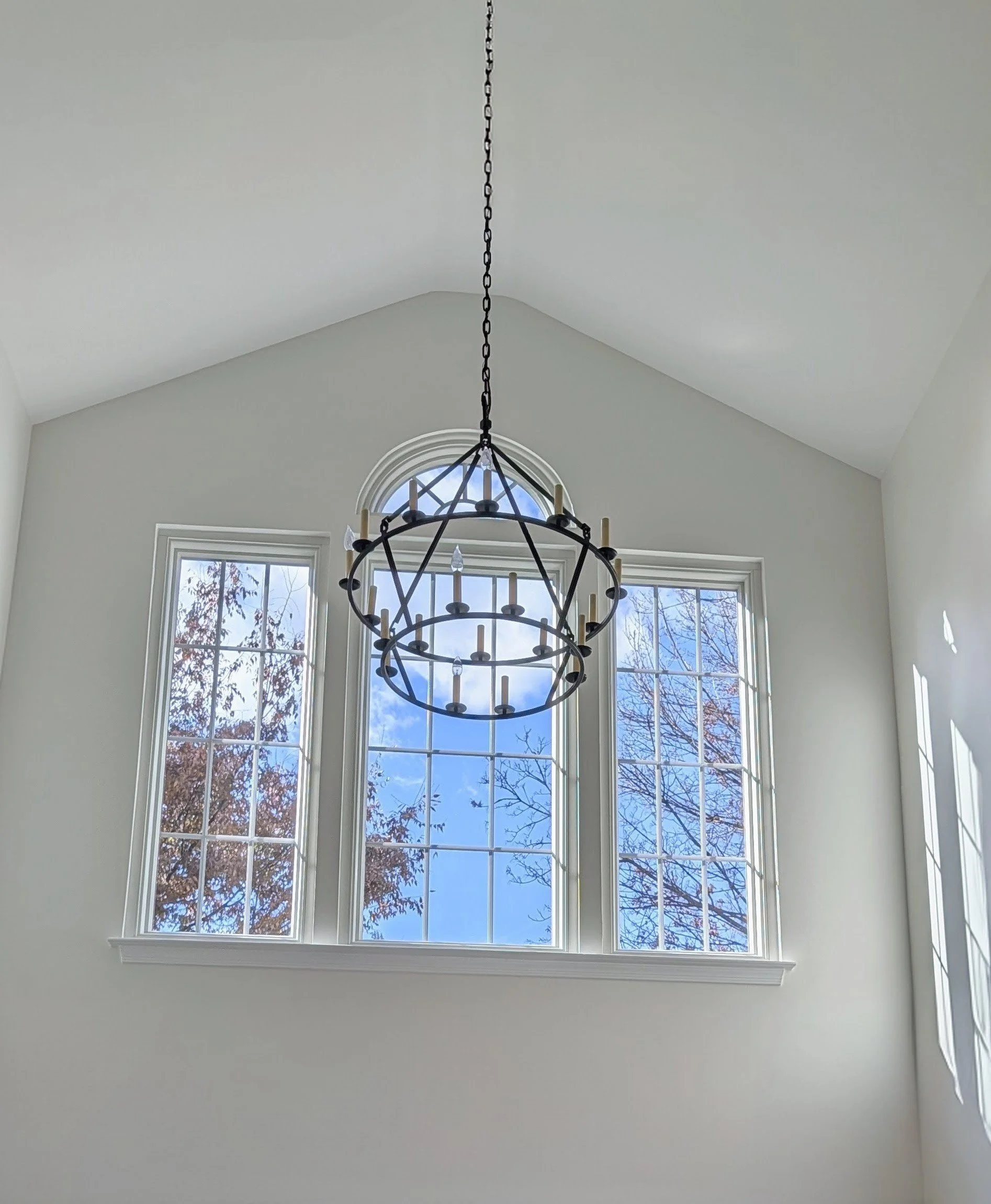Bright room with white walls and three large windows showing blue sky and trees outside, and a black metal chandelier hanging from the ceiling.