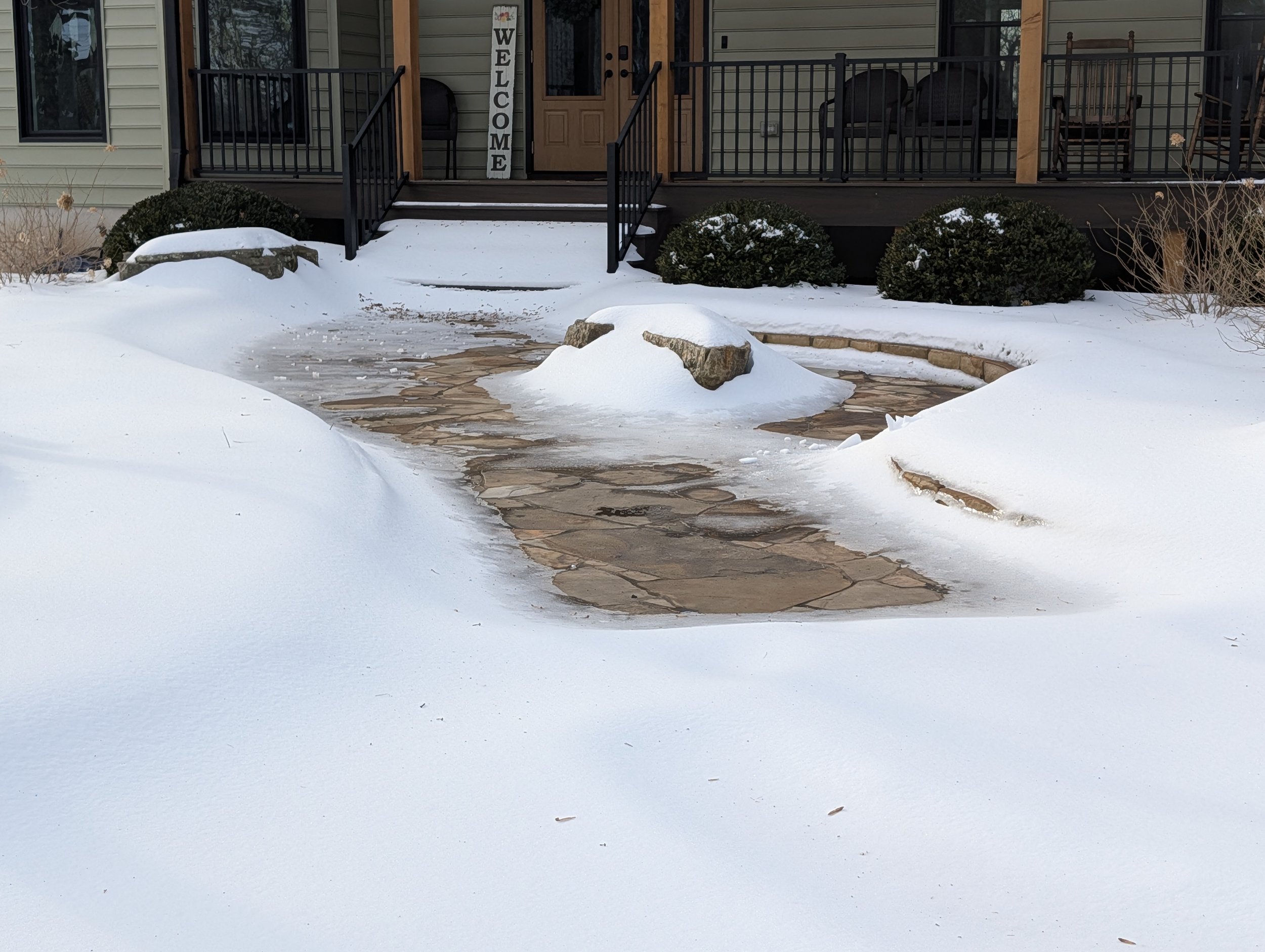Snow-covered sidewalk leading up to a house with a porch, a welcome sign on the door, and bushes around the entrance, during winter.