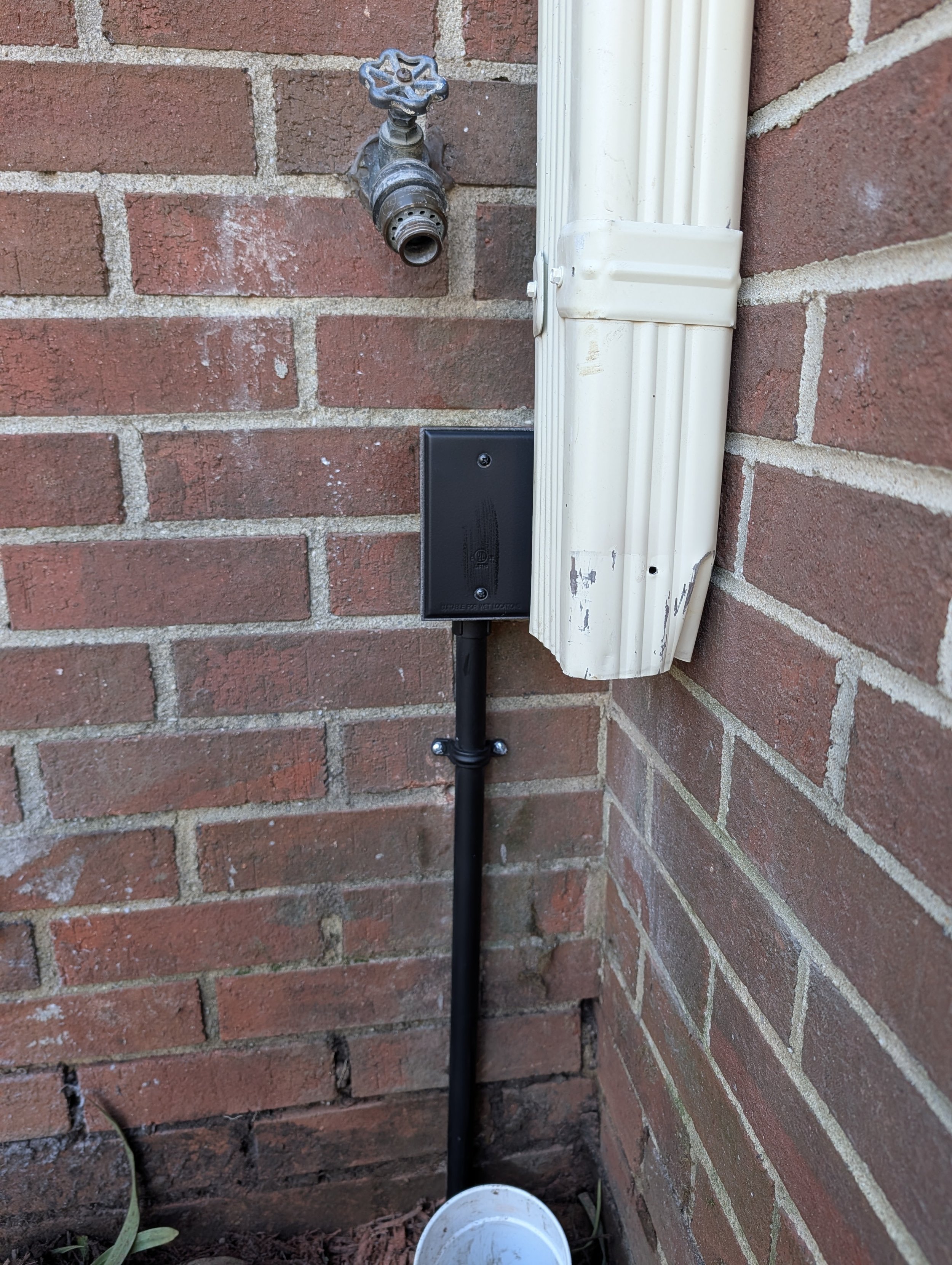 An outdoor water faucet with a nozzle, a black electrical box, and a black pipe going down to a white bucket, against a reddish brick wall.