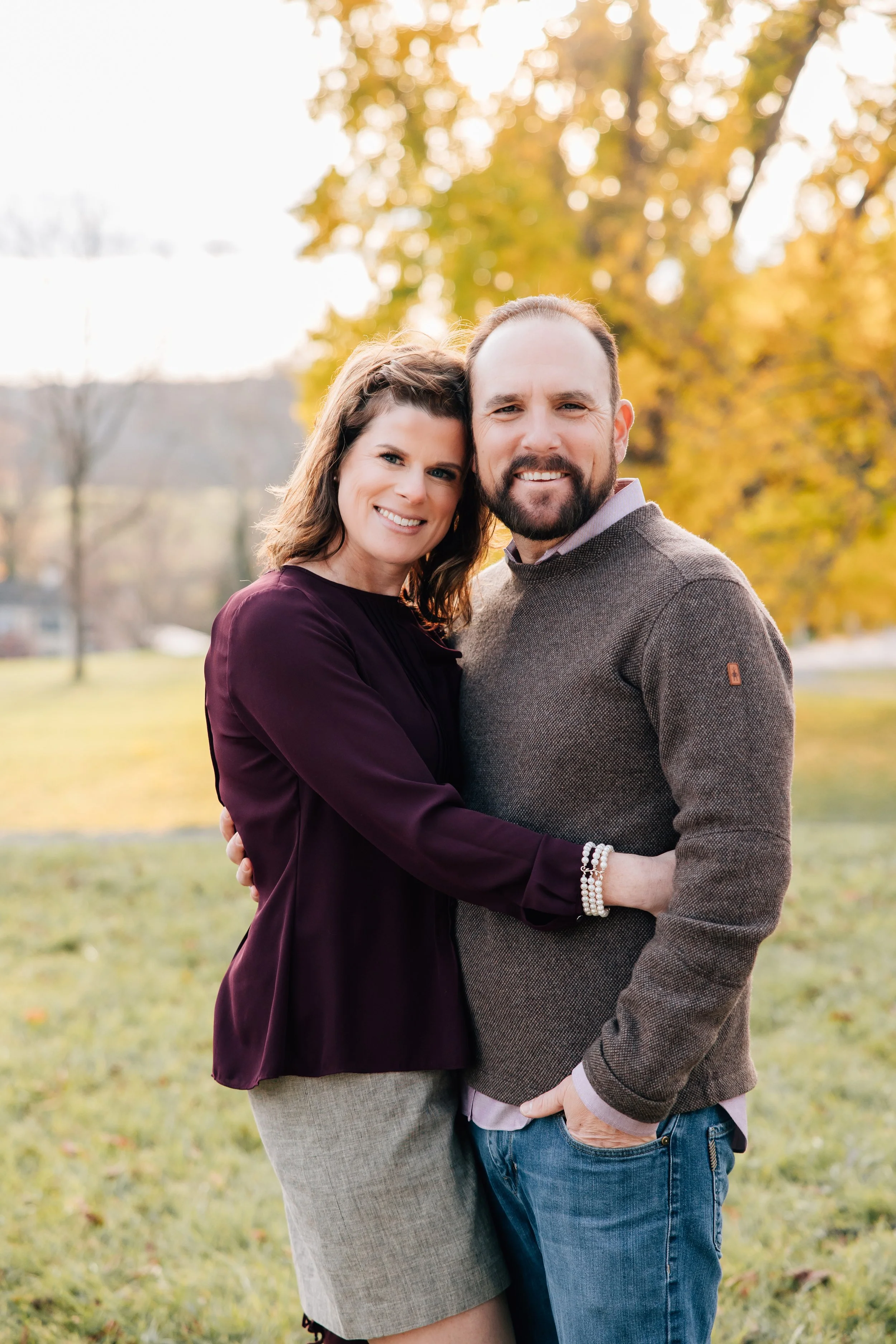A happy couple is standing close together outdoors during fall, smiling at the camera, with trees showing yellow and orange leaves in the background.