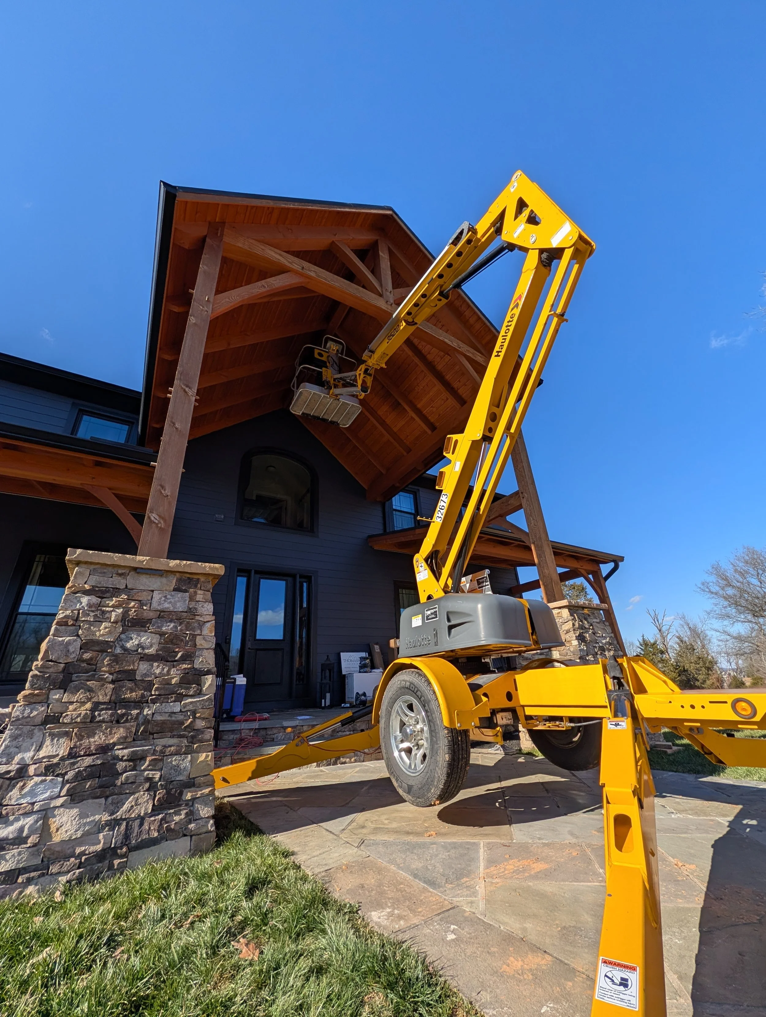 A yellow hydraulic lift tower is extended to reach the roof of a house, which has a wooden porch and stone columns, in a clear sunny day.