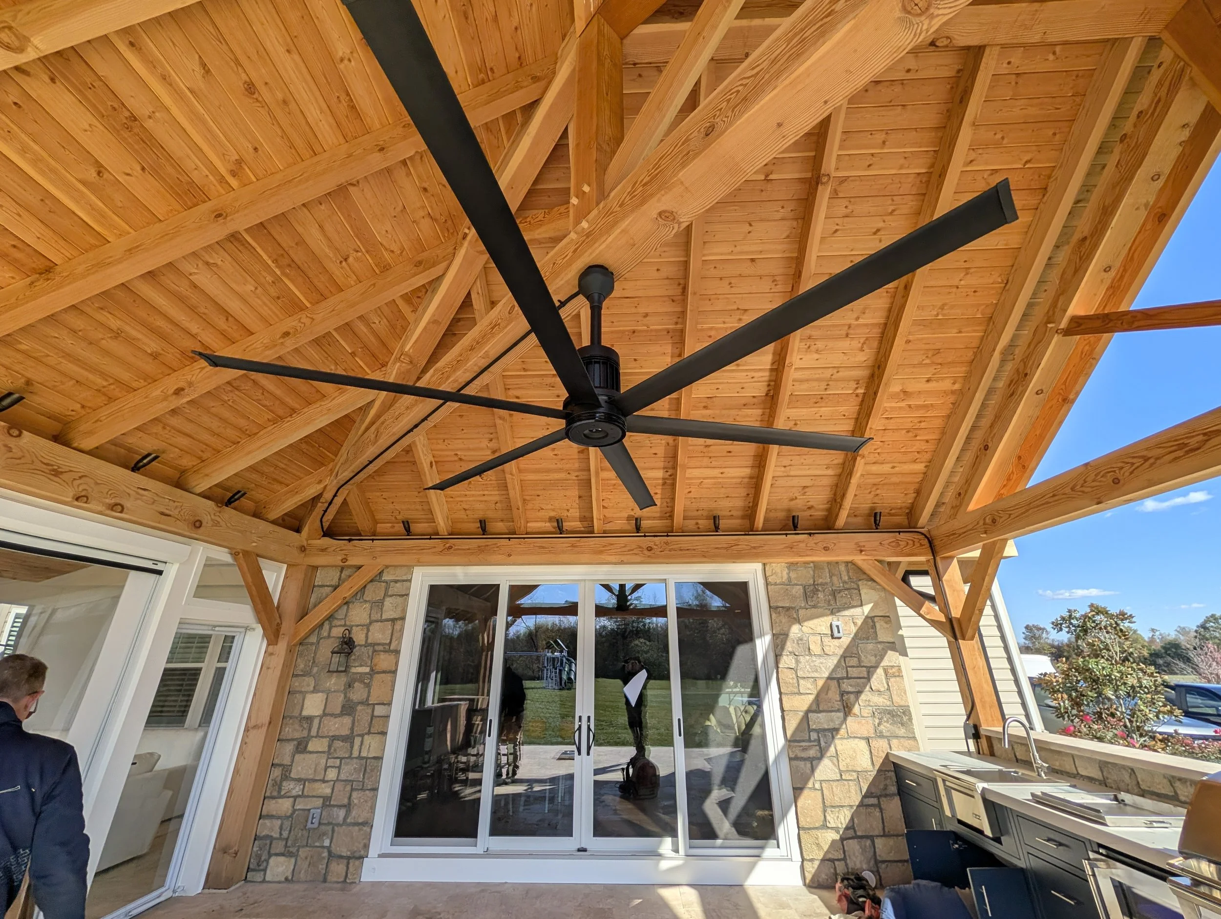 Covered outdoor patio with wooden ceiling, black ceiling fan, stone wall, sliding glass door, and outdoor kitchen sink.