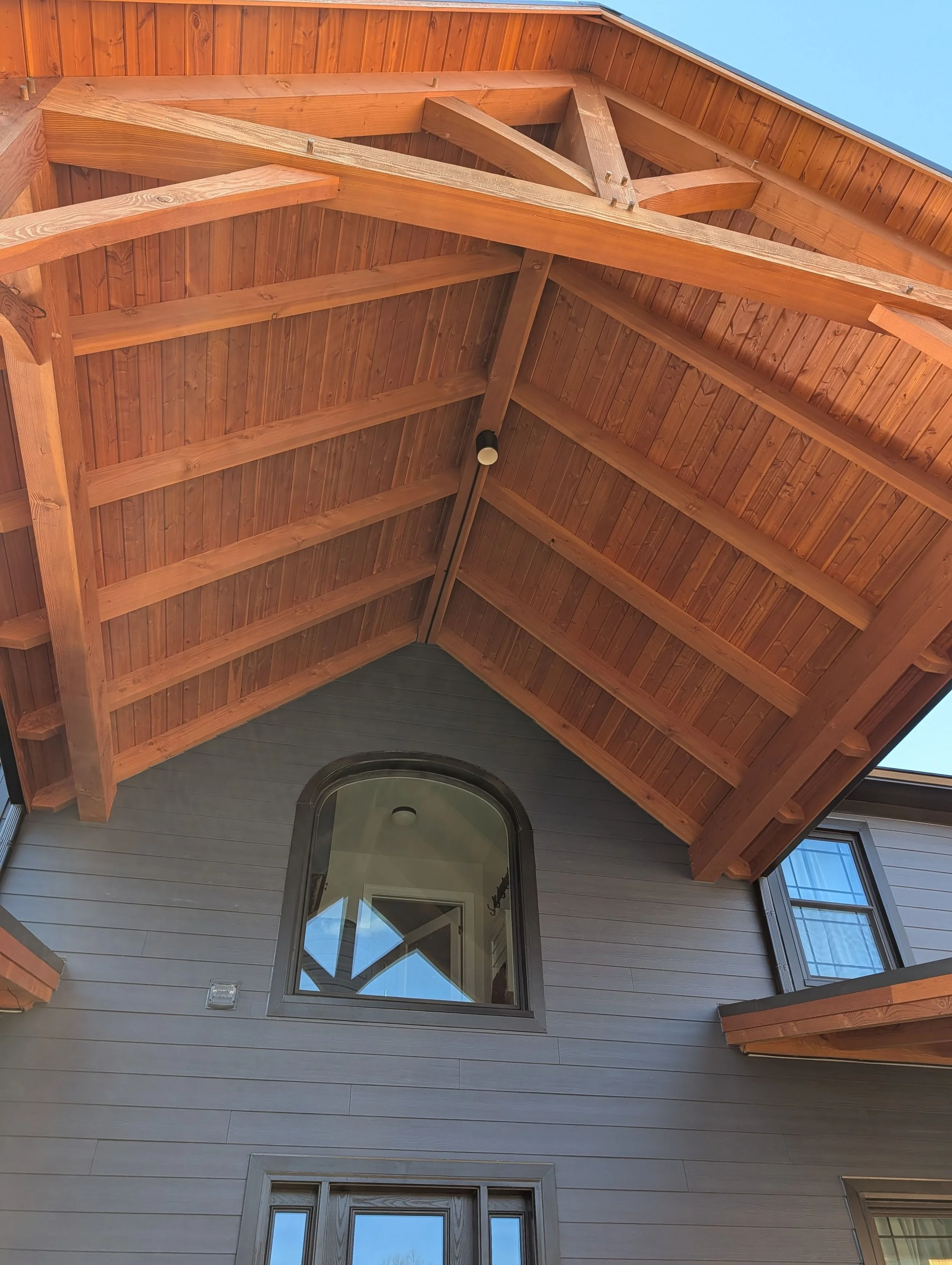 Looking up at a house's wooden roof overhang, showing the underside of the wooden beams and shingles, with a large arched window on the second floor and a door below.