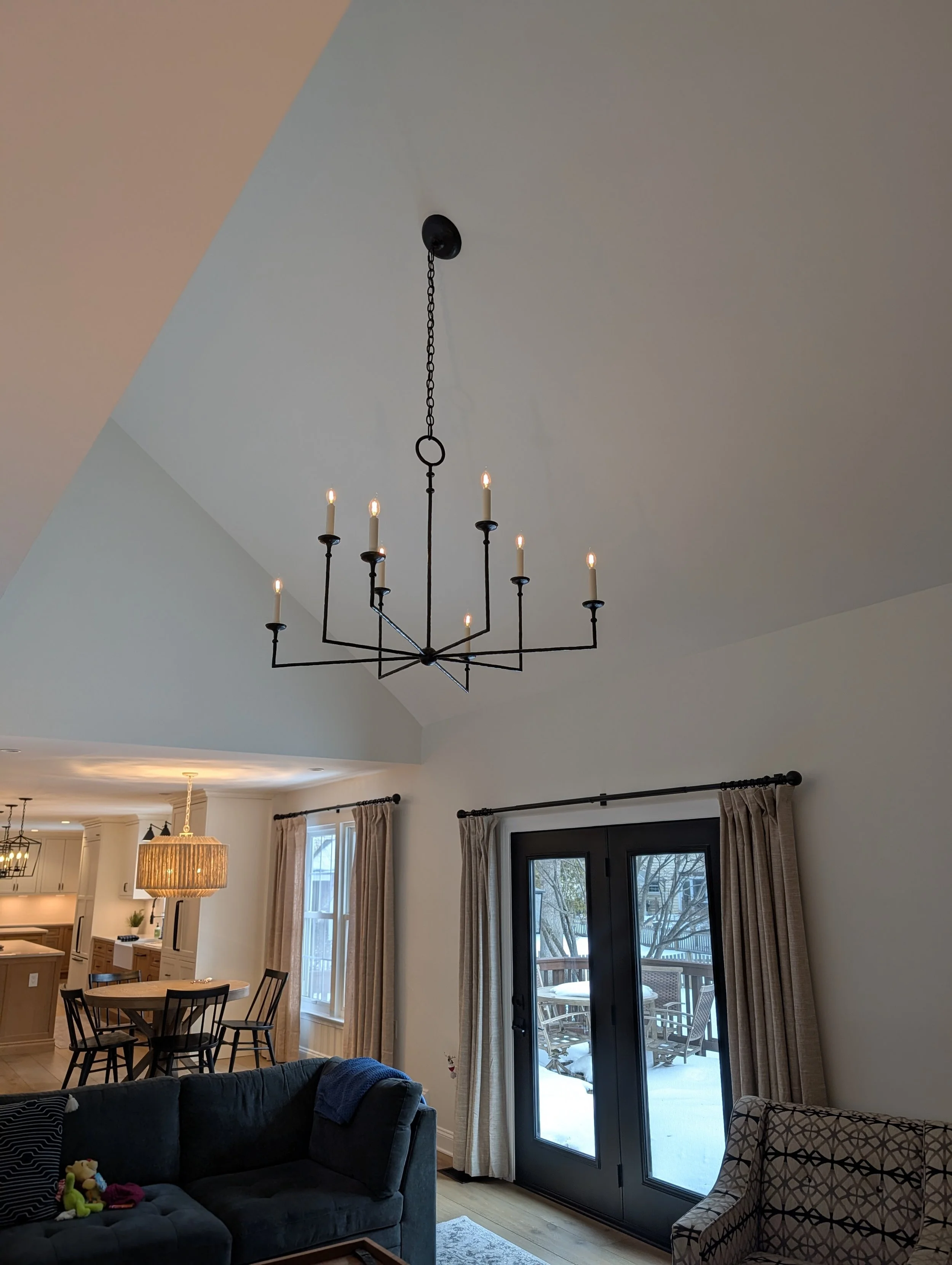 Living room with black chandelier, beige curtains, glass patio door showing outdoor snow, sofa, patterned armchair, and dining area in background.