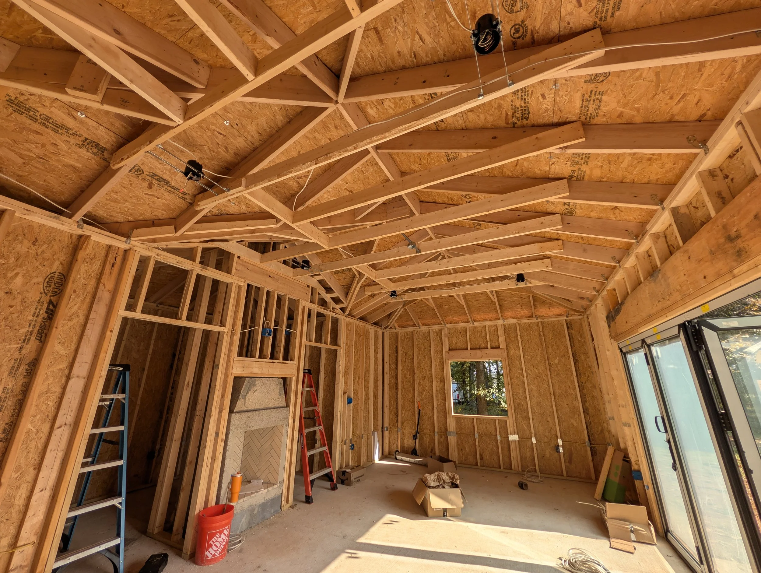 Interior of a house under construction, showing wooden framing, an unfinished fireplace, a ladder, and construction tools, with sunlight coming through the windows.