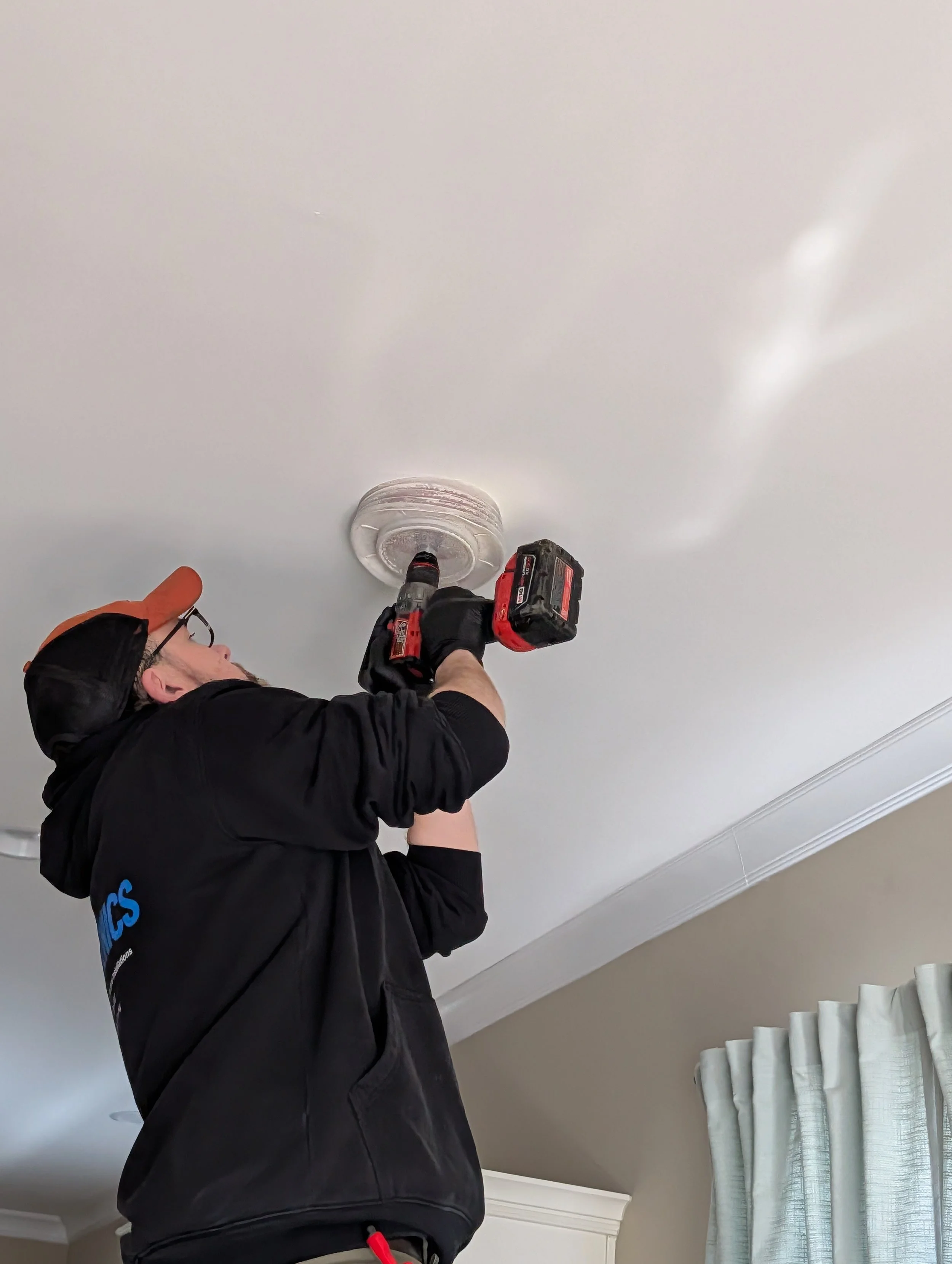 A man wearing an orange baseball cap and black hoodie installing or repairing a ceiling smoke detector with a cordless drill.