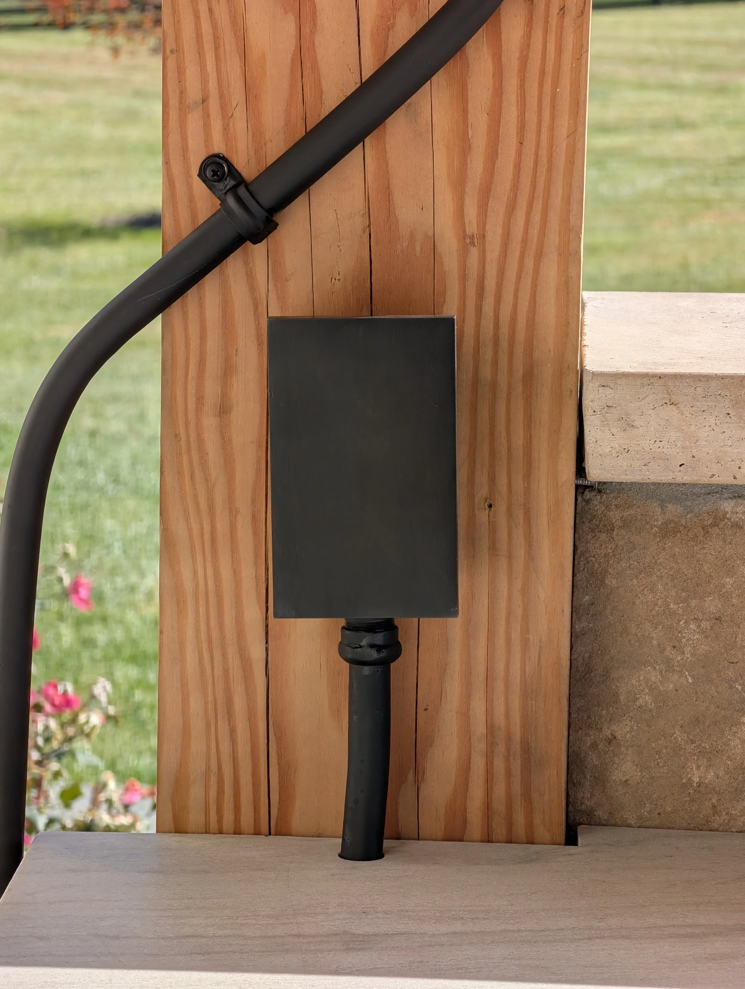 Close-up view of an electrical junction box and conduit attached to a wooden post, with a black metal pipe running downward, outside structure with a grassy area and some flowers in the background.