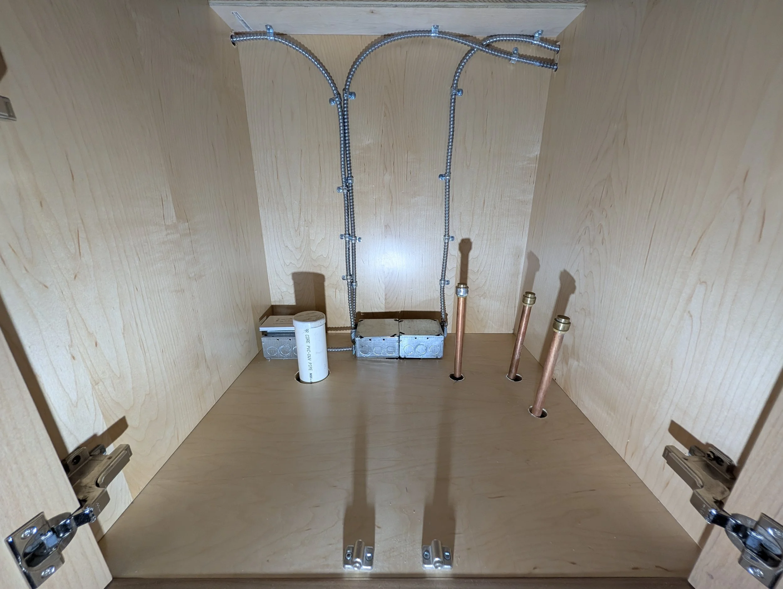 Interior of a wooden cabinet with electrical wiring, metal conduit, copper pipes, and a roll of pipe thread tape.