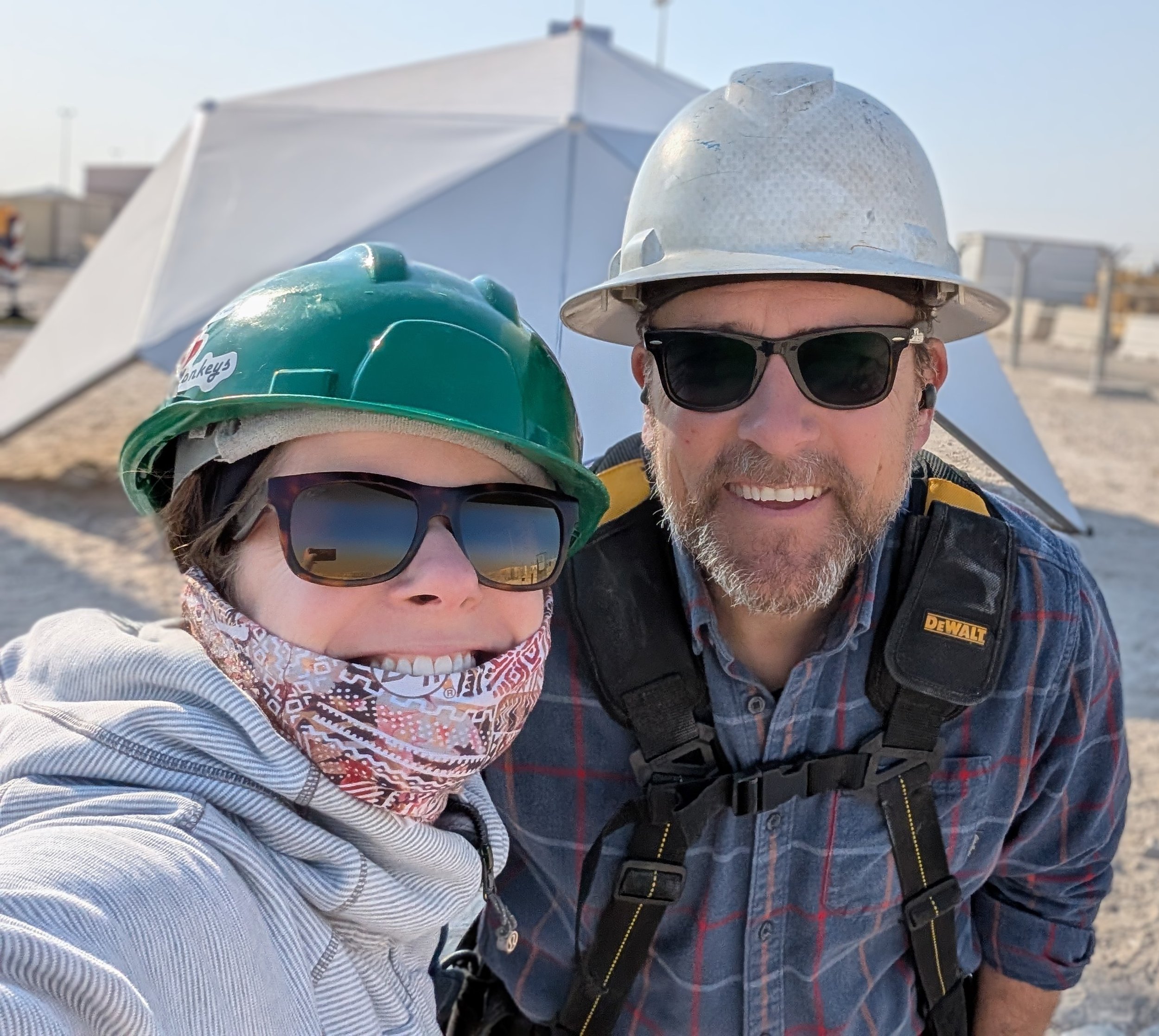 A woman and a man in safety gear smiling at the camera, with tents and a construction site in the background.