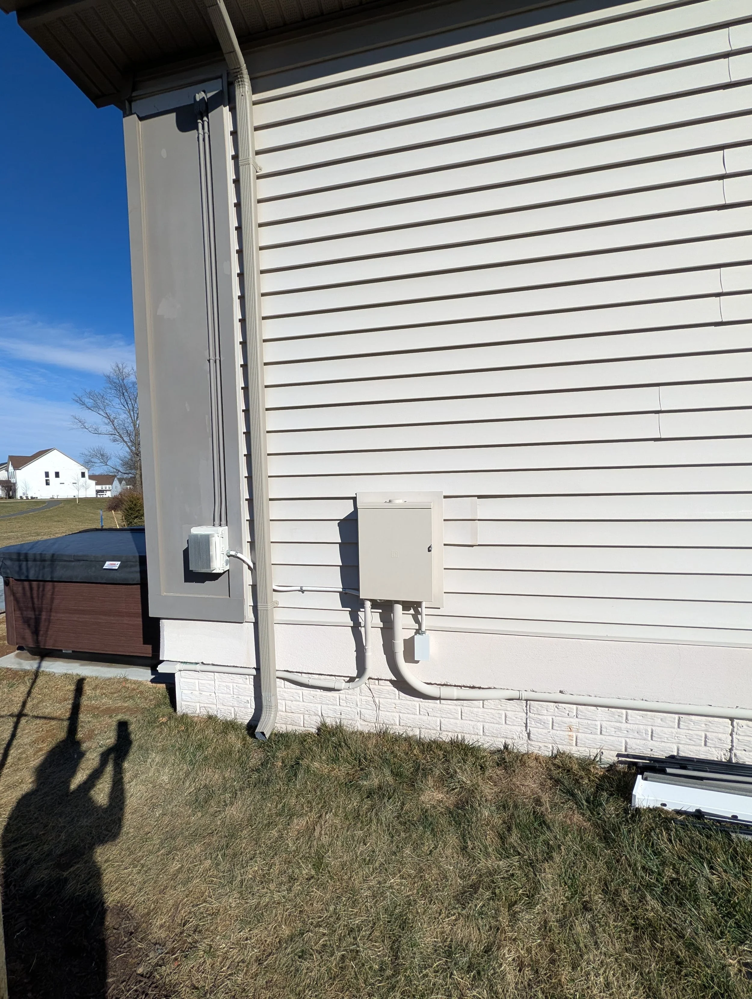 Exterior of a house with white vinyl siding, showing electrical panels, conduit pipes, and a grassy yard with shadows of two people taking the photo.