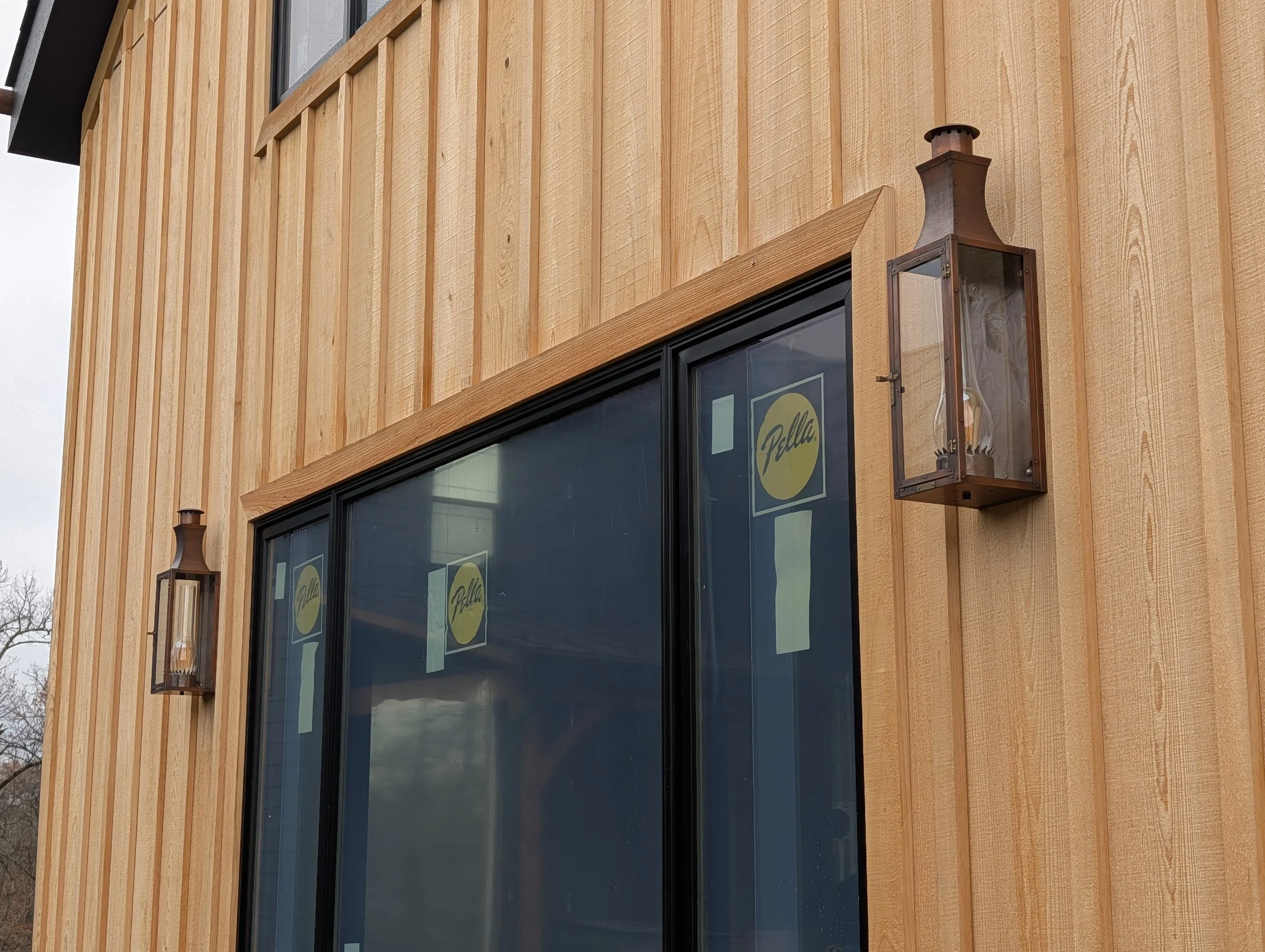 Close-up of an exterior wooden wall with large window, two vintage-style lanterns, and yellow Pella stickers on the window.