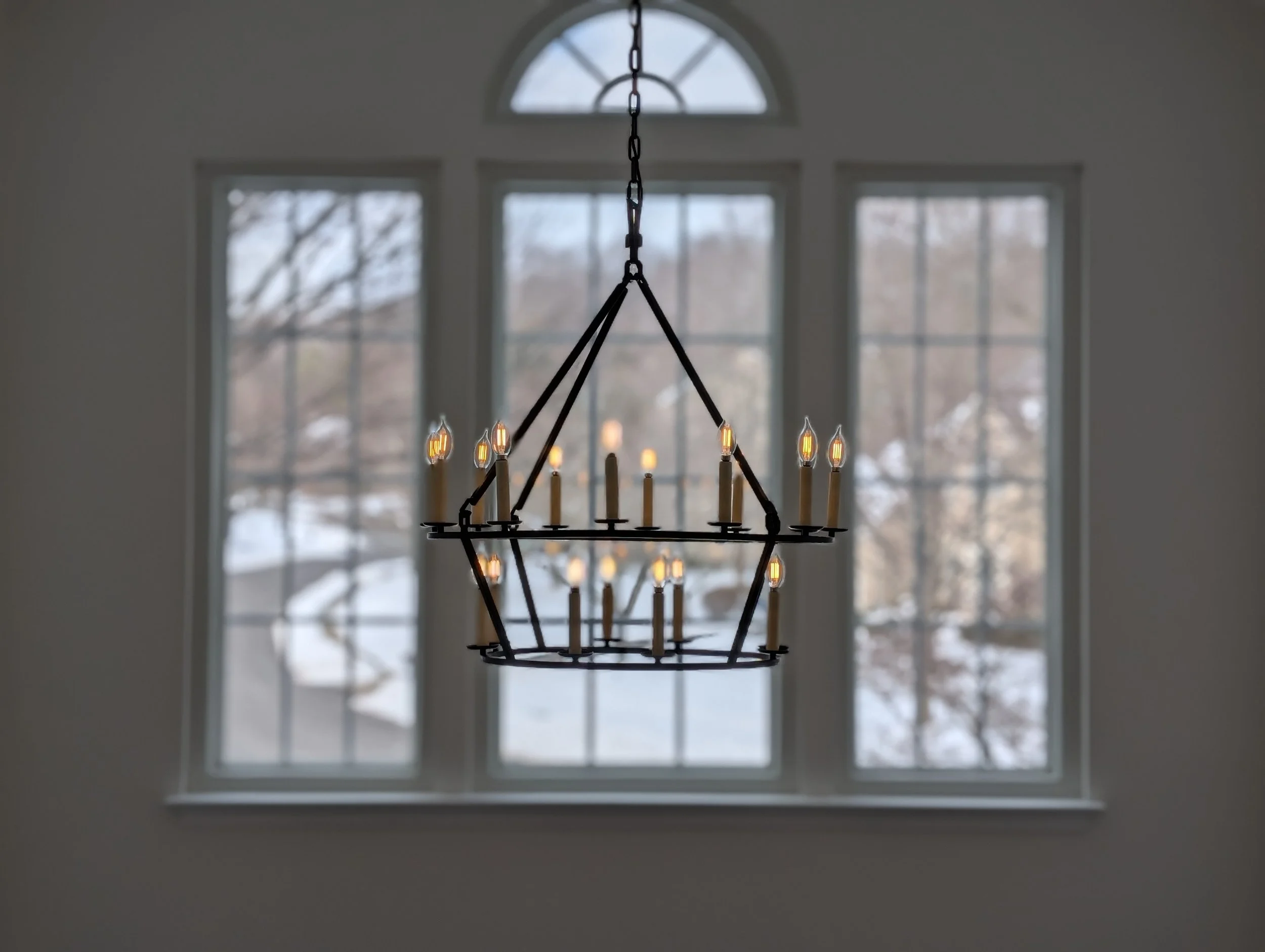 Chandelier with lit bulbs hanging in front of a large window showing a snowy outdoor scene.