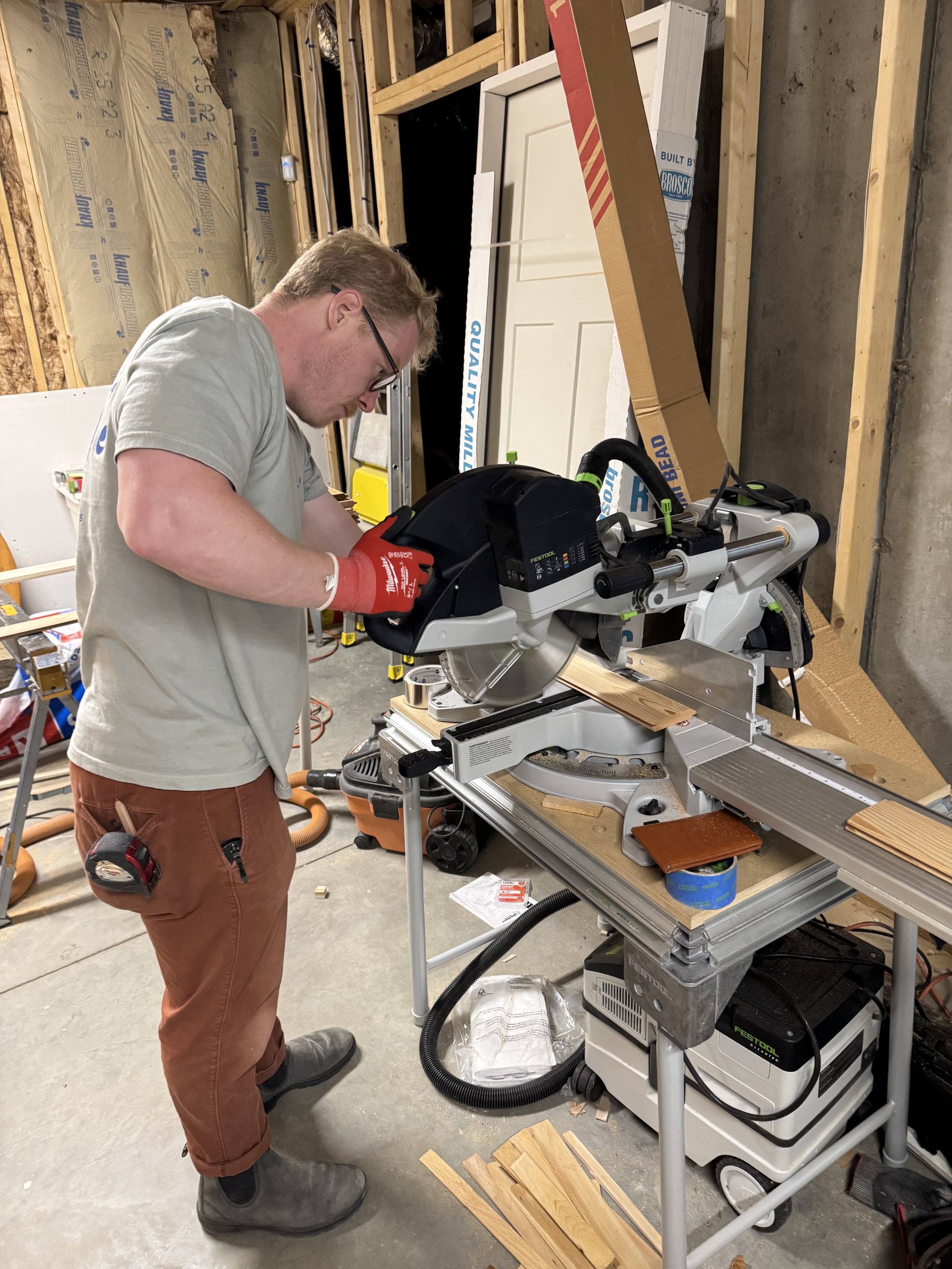 A man wearing glasses and red gloves operating a miter saw in a workshop, cutting a piece of wood.