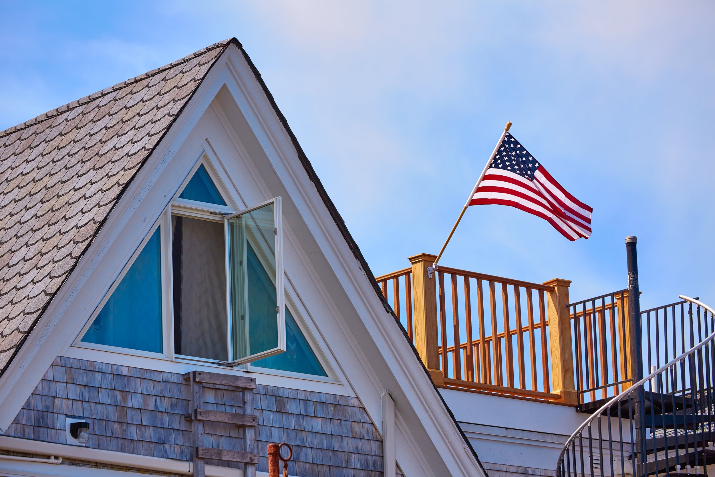 A house with a triangular window and a rooftop deck with an American flag on a pole. Blue sky in the background.