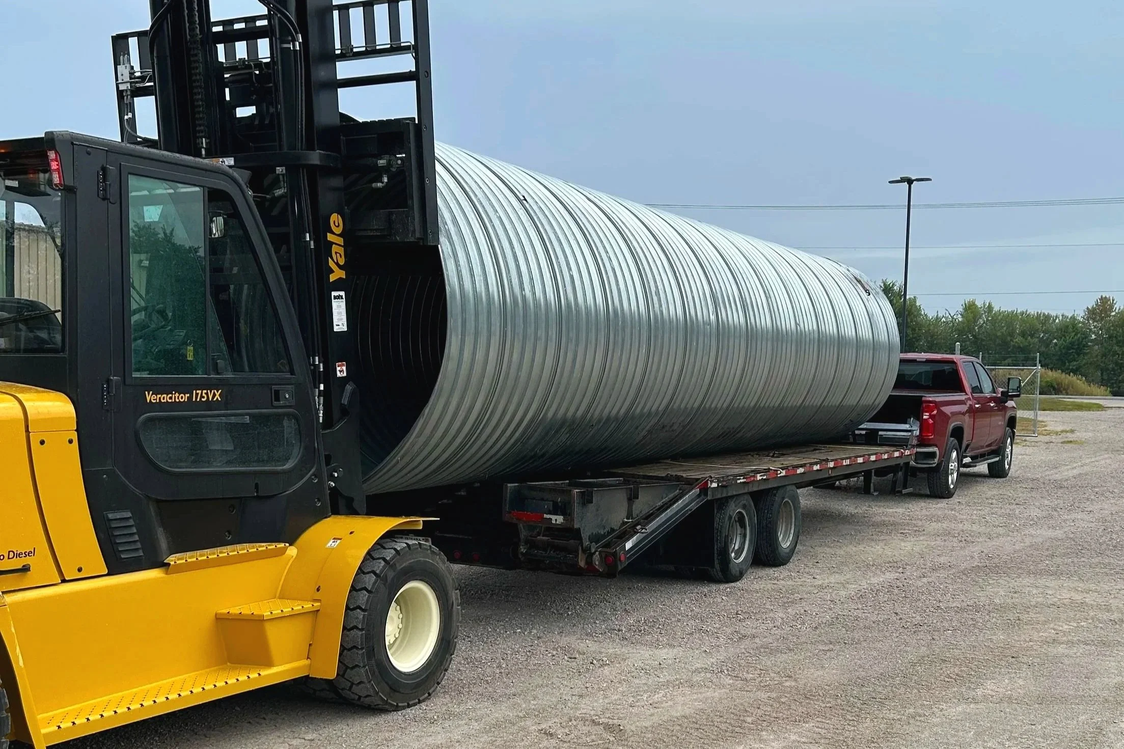A yellow forklift carrying a large metal pipe on a flatbed truck in an open lot.