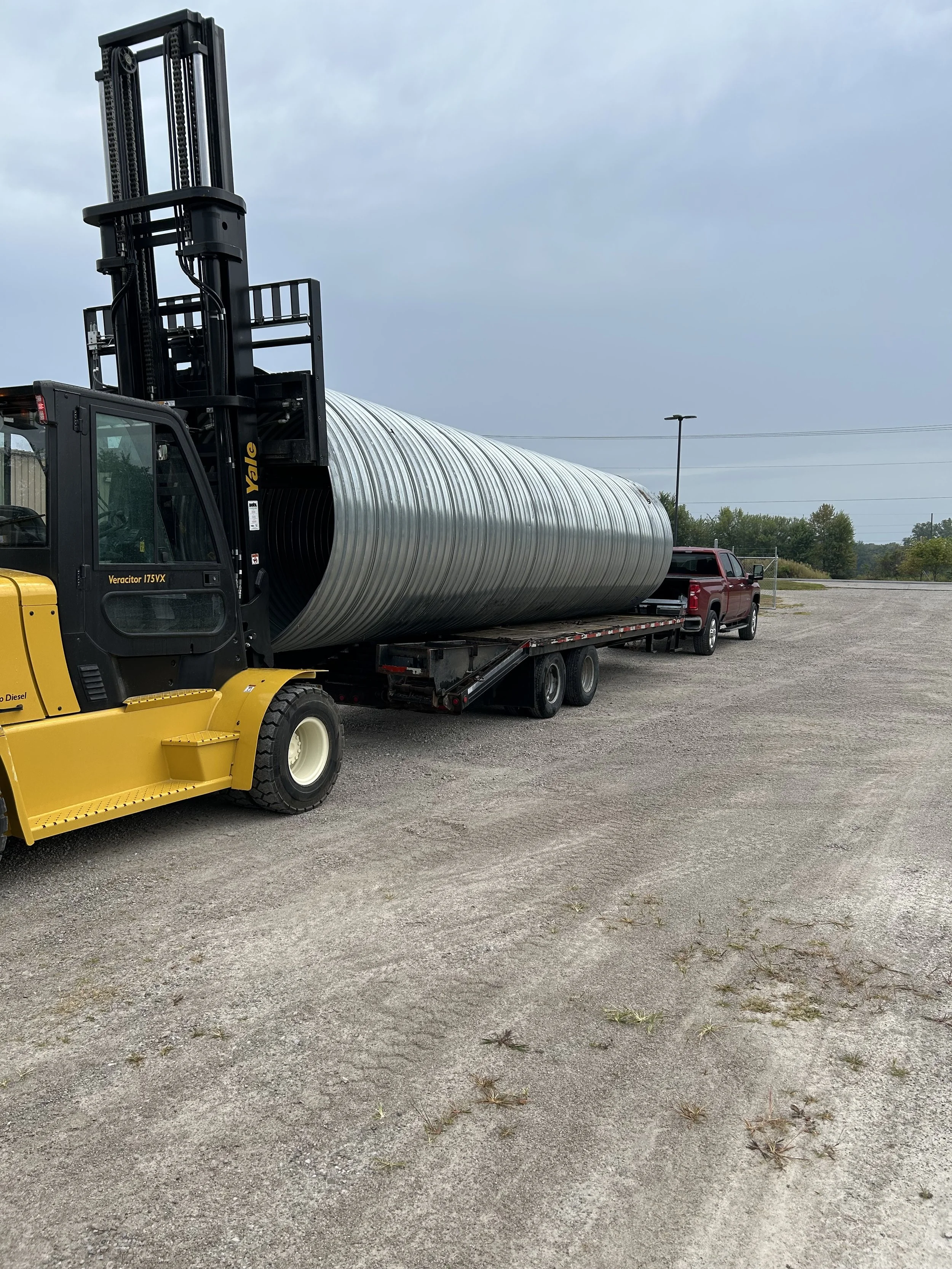 A forklift vehicle lifting a large metallic cylindrical storage tank onto a flatbed truck at an outdoor lot.