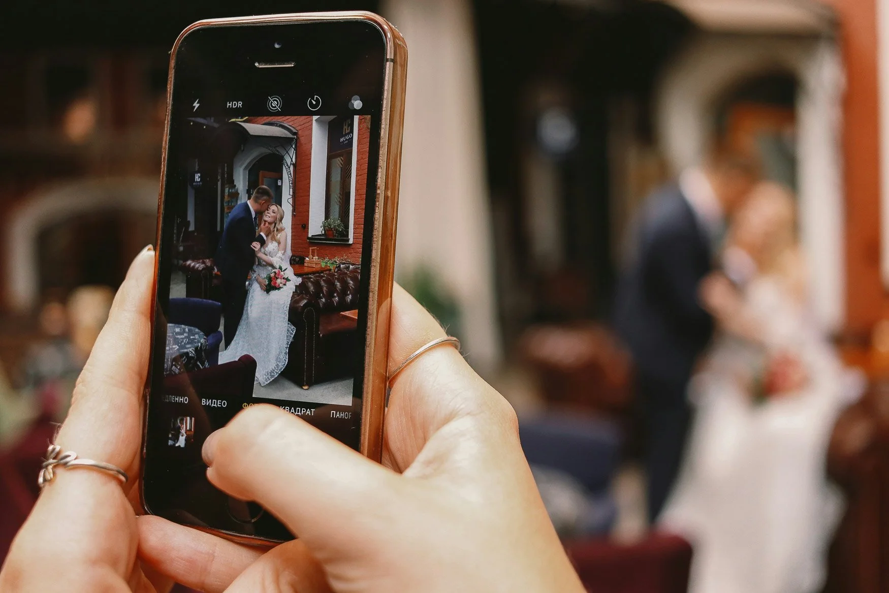 A guest photographs a bride and groom with an iPhone at a wedding, the couple visible both on the phone screen and blurred in the background.
