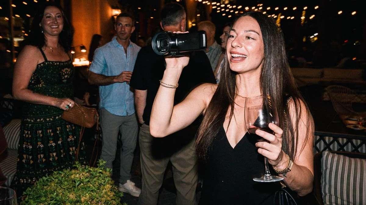 A woman in a black dress smiles while holding a camcorder to her eye and a glass of red wine at an outdoor evening event with guests mingling behind her.