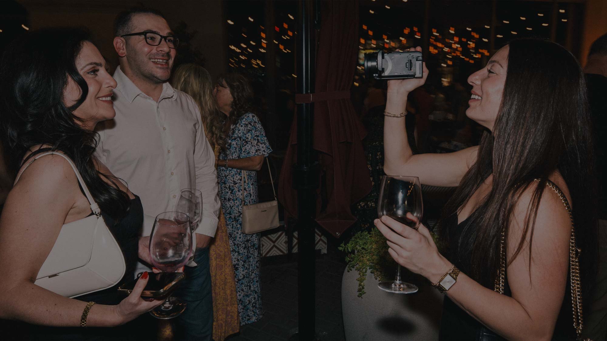 A smiling woman points a camcorder at two guests holding wine glasses at a dimly lit evening event, documenting real, unfiltered moments the way only a camcorder can.