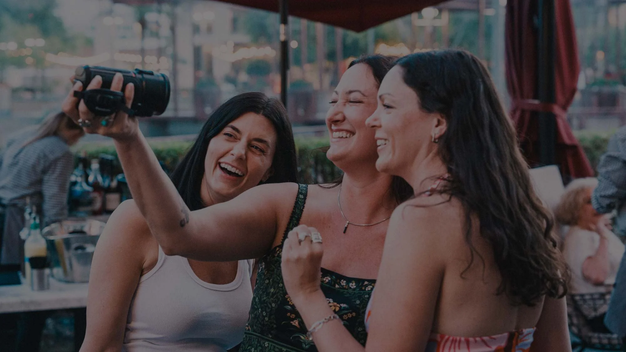 Three women laugh together while taking a selfie with a camcorder at an outdoor evening event, with string lights and a patio bar visible in the background.